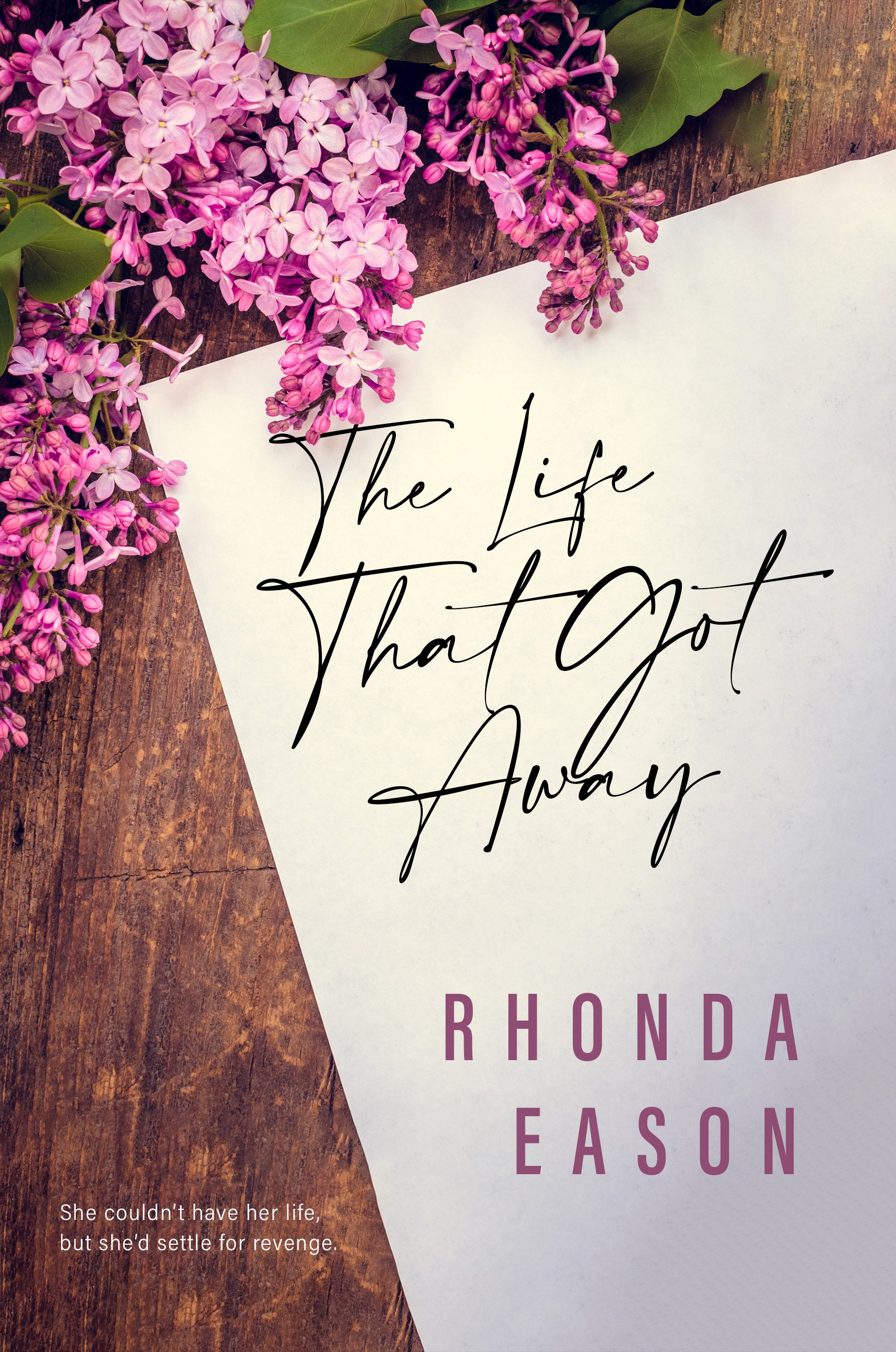 Close-up of pink lilac flowers on a wooden surface with a white card displaying handwritten text 'The Life That Got Away' and partially visible text by author 'RHONDA EASON'.