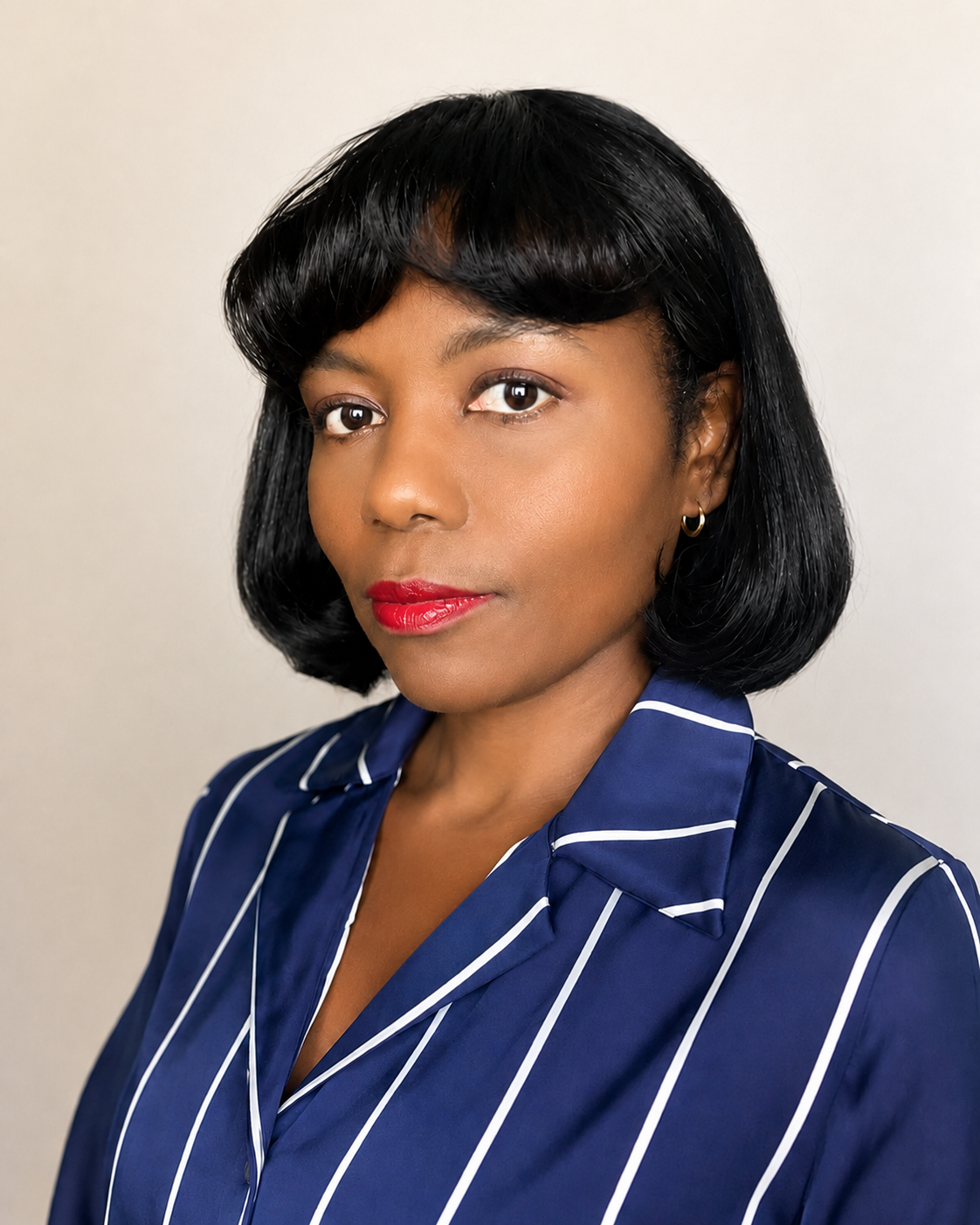 A woman with black hair in a bob style, wearing a blue and white striped collared shirt, red lipstick, and gold hoop earrings, standing against a plain background.