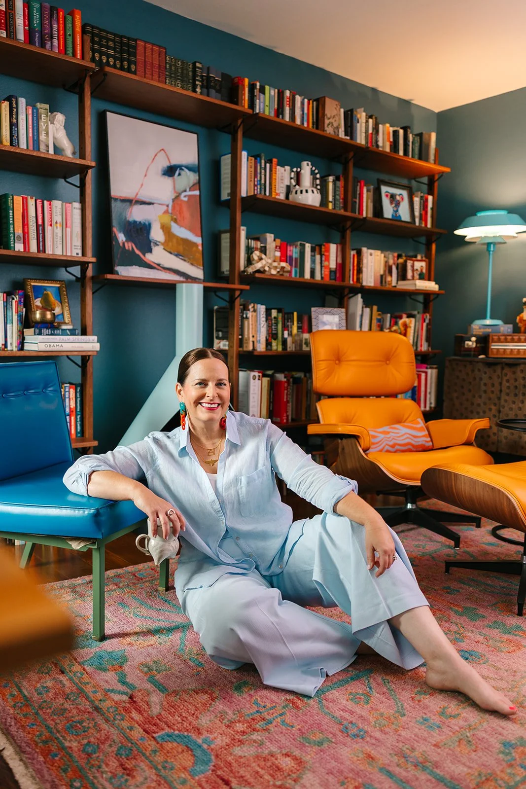 A woman with brown hair sitting on the floor in a cozy living room, smiling. She is dressed in a white pajama set, with earrings and necklaces. Behind her, there is a large bookshelf filled with colorful books, a modern orange chair, and a blue armchair. The room has a warm, inviting atmosphere with a patterned rug and a blue wall.