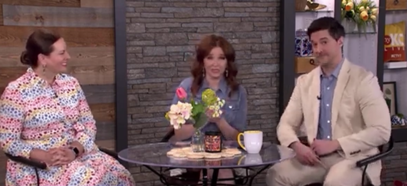 Three people sitting around a round table on a TV show set. The woman on the left is wearing a colorful patterned dress, the woman in the middle is holding flowers, and the man on the right is wearing a beige suit. There are flowers, a mug, and a jar on the table.