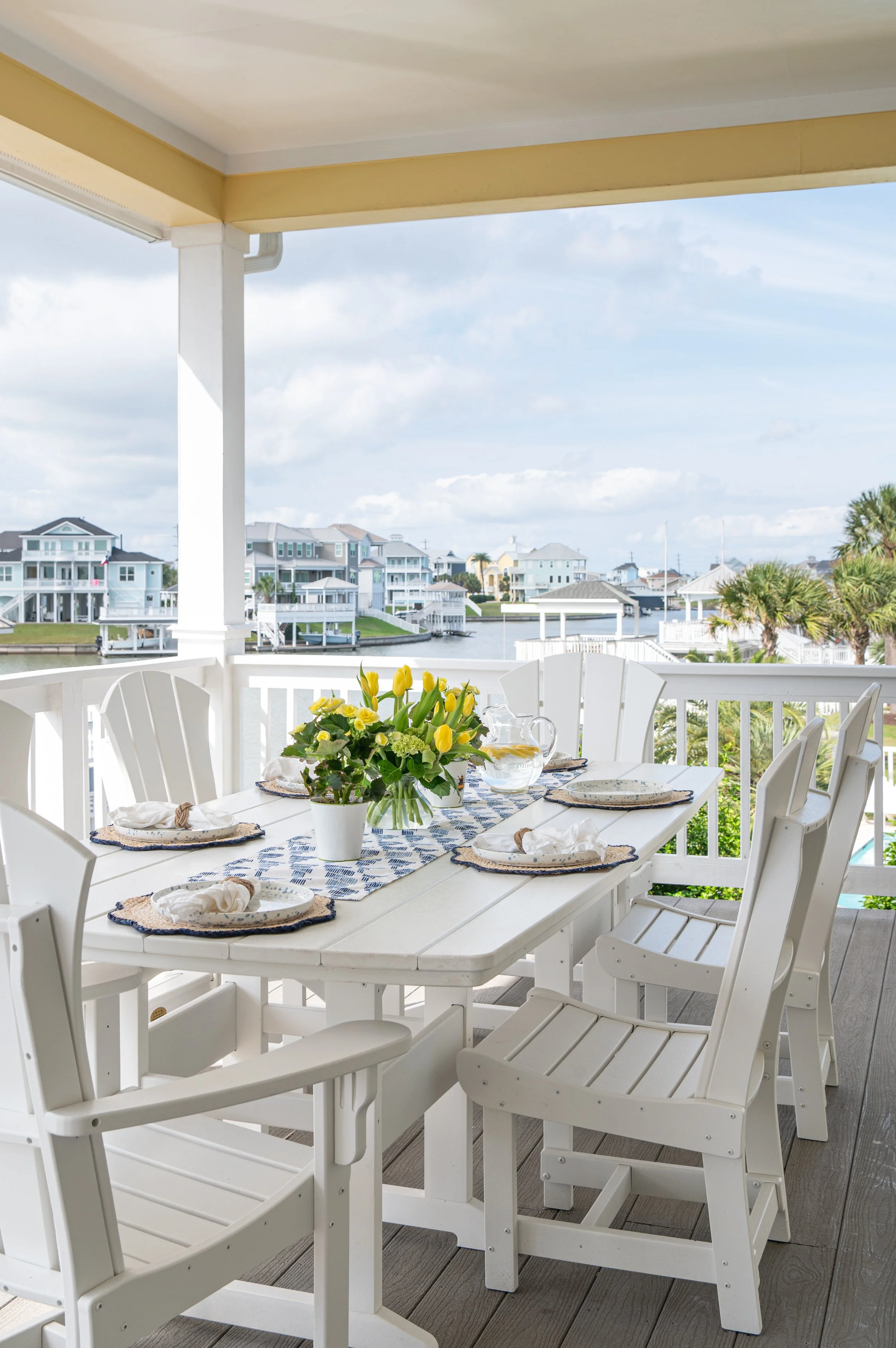 A balcony dining area with a white table set with plates, napkins, and a flower arrangement with yellow tulips, overlooking houses and water in a coastal neighborhood.
