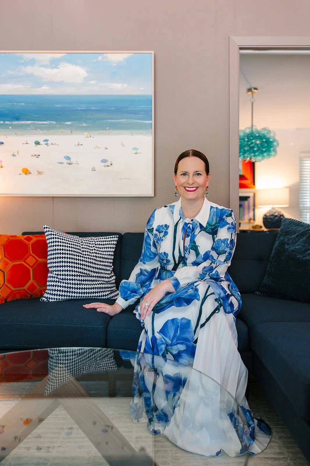 A woman with brown hair and red lipstick sitting on a dark sofa in a living room, wearing a white dress with blue floral patterns, with colorful pillows and a large beach scene painting behind her.
