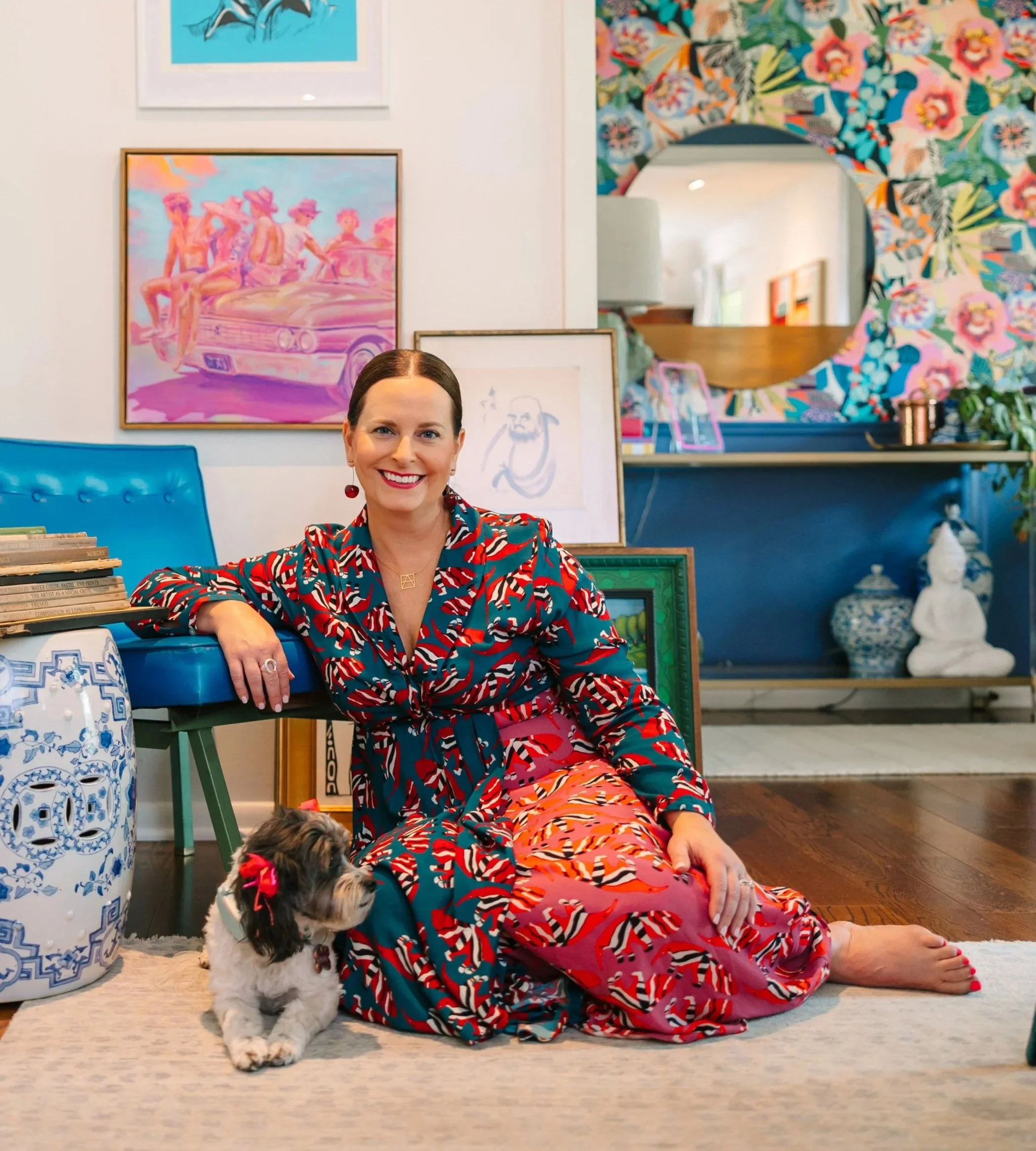 A woman with dark hair in a bun smiling while sitting on the floor in a room decorated with colorful artwork and patterned wallpaper. She is wearing a vibrant dress with red, blue, and orange patterns. A small dog with black, white, and brown fur and a red bow on its head is sitting next to her. There are shelves with framed pictures and decorative items behind her.