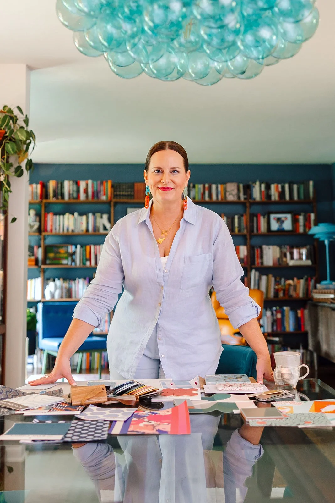 A woman in a white shirt standing behind a table with fabric samples and color swatches, in a room with a large bookshelf in the background and a chandelier overhead.