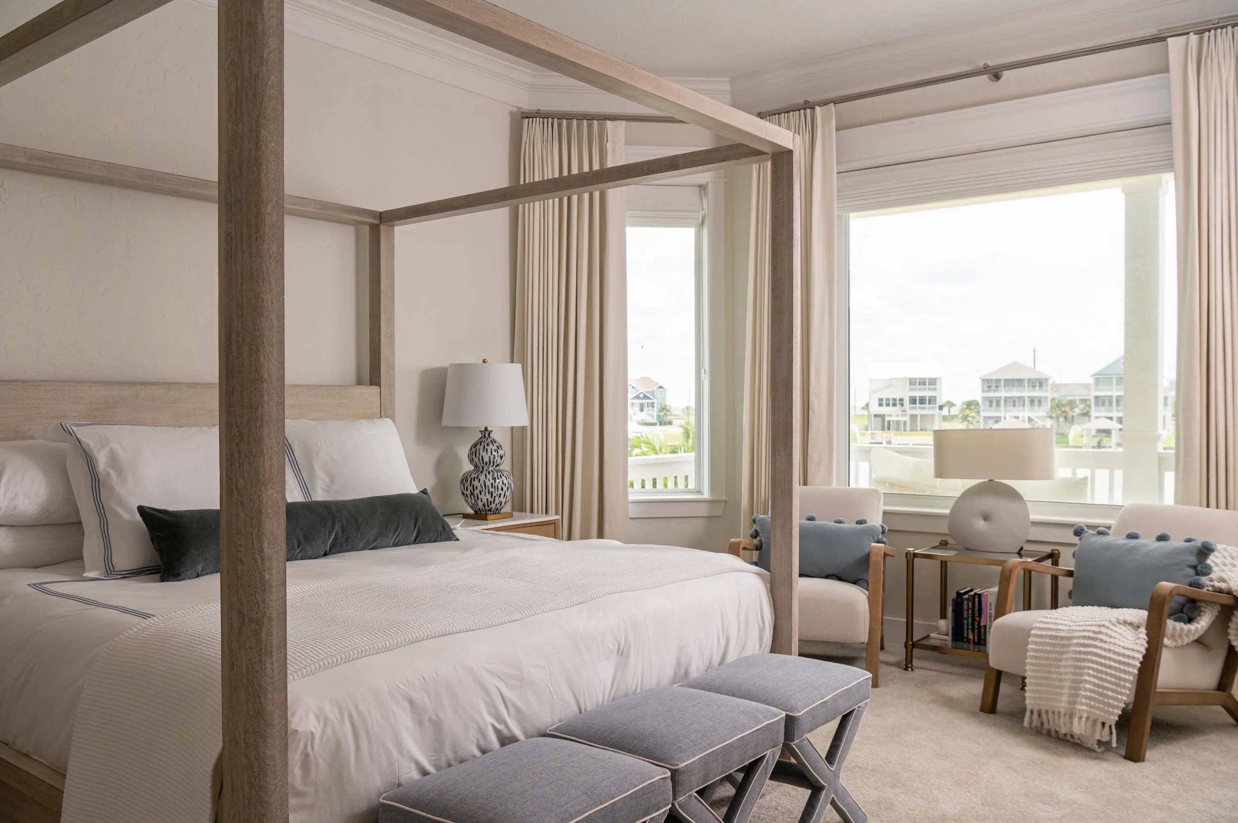 A cozy bedroom with a white bed, plush pillows, and a wooden canopy frame. Large windows with cream curtains reveal an outdoor view of houses and greenery. The room features a bedside lamp, decorative vases, and seating areas with a woven basket and small upholstered stools.