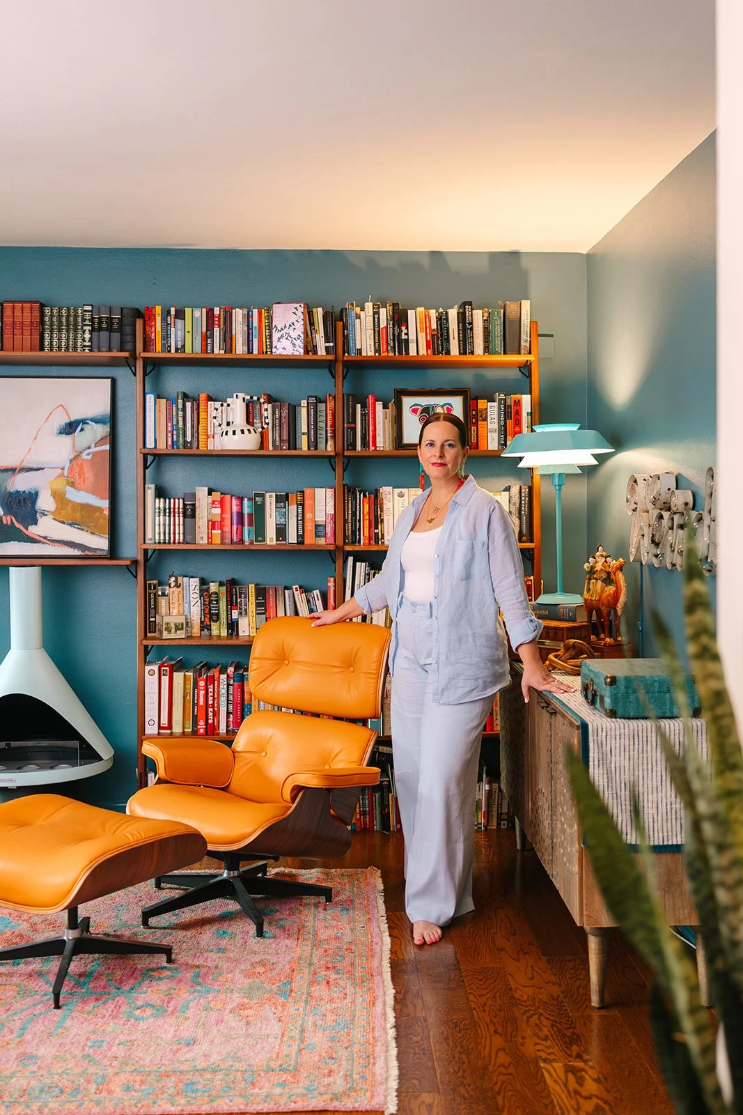A woman standing in a cozy, well-decorated living room with a blue wall, wooden bookshelf filled with books, a colorful abstract painting, a modern orange lounge chair and ottoman, a patterned area rug, and a side table with decorative items and a turquoise lamp.