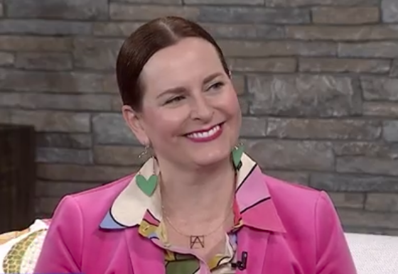 A smiling woman with red hair, wearing a pink blazer and colorful earrings, seated against a stone wall backdrop.