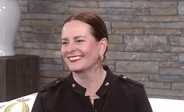 A woman with red hair and earrings, smiling, sitting against a brick wall with a sofa and cushion.