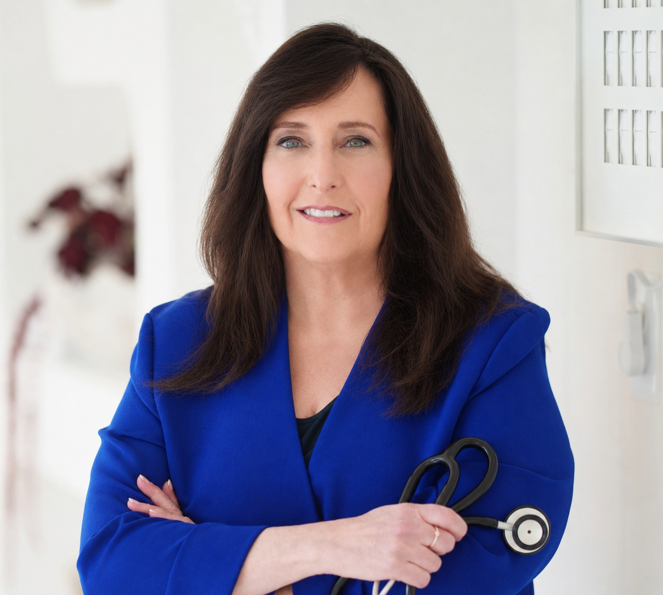 A female healthcare professional with long brown hair, wearing a bright blue blazer, holding a stethoscope, standing in a clinical setting.