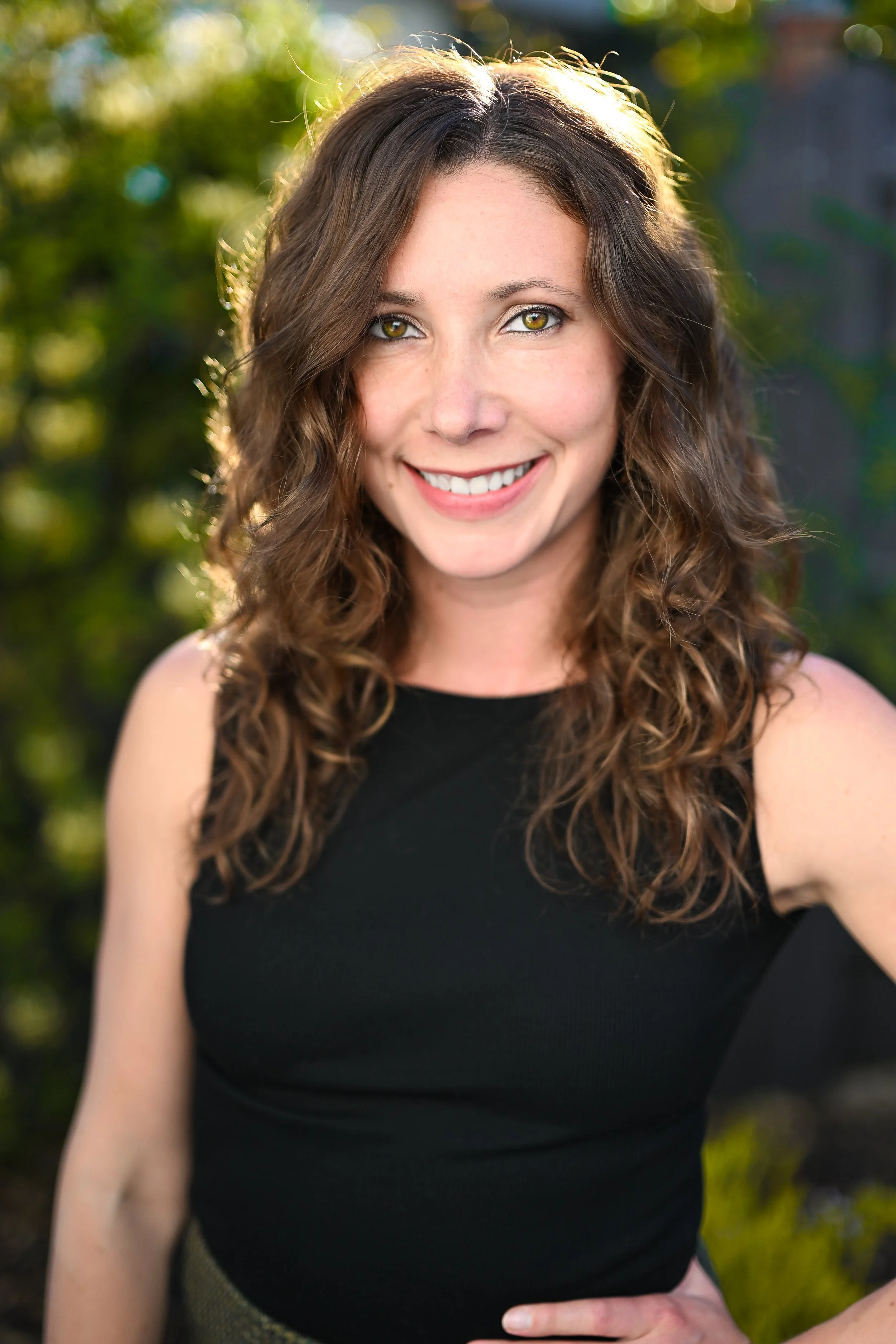 A woman with curly brown hair and hazel eyes smiling outdoors in sunlight, wearing a black sleeveless top.
