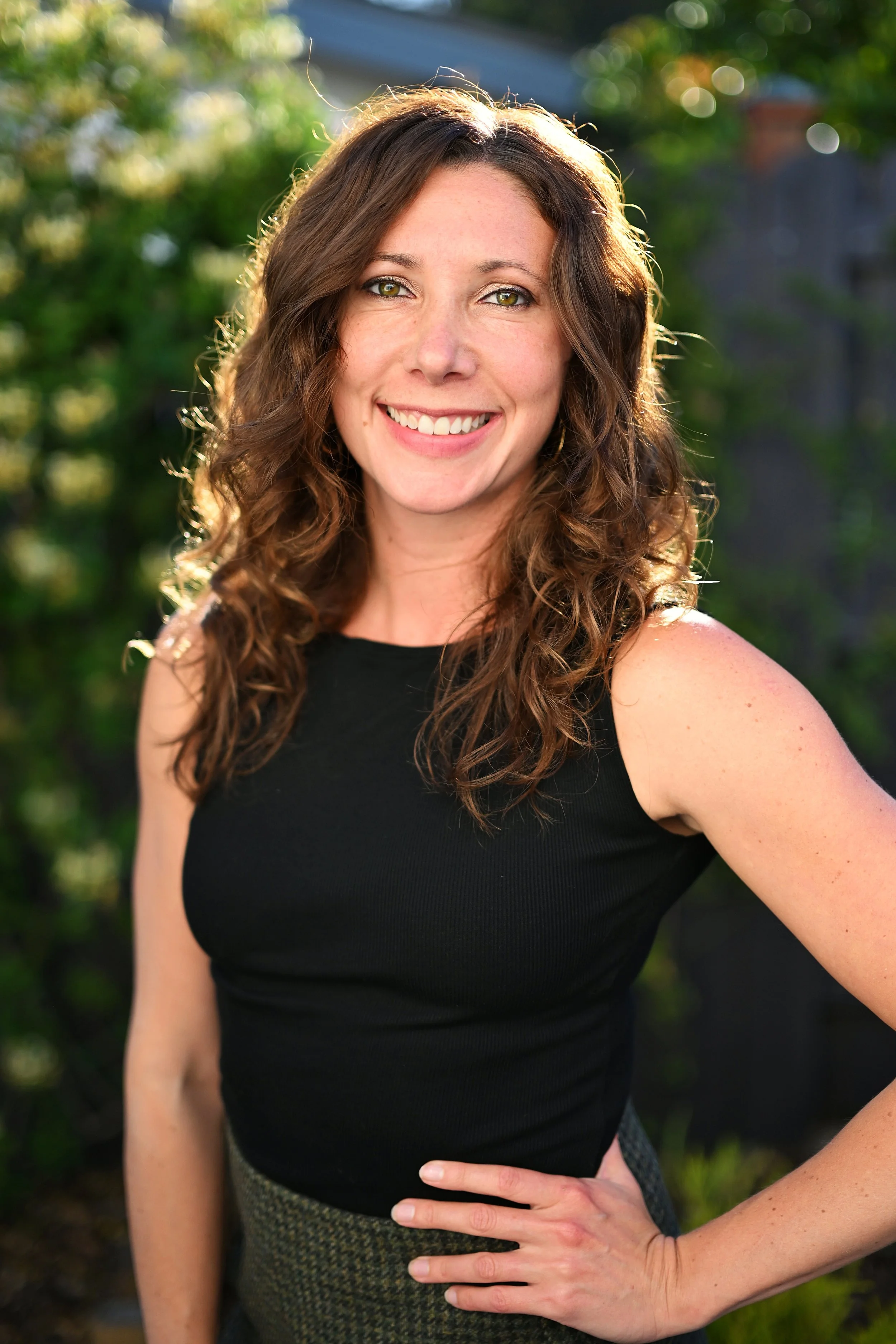A woman with curly brown hair and hazel eyes smiling outdoors on a sunny day.