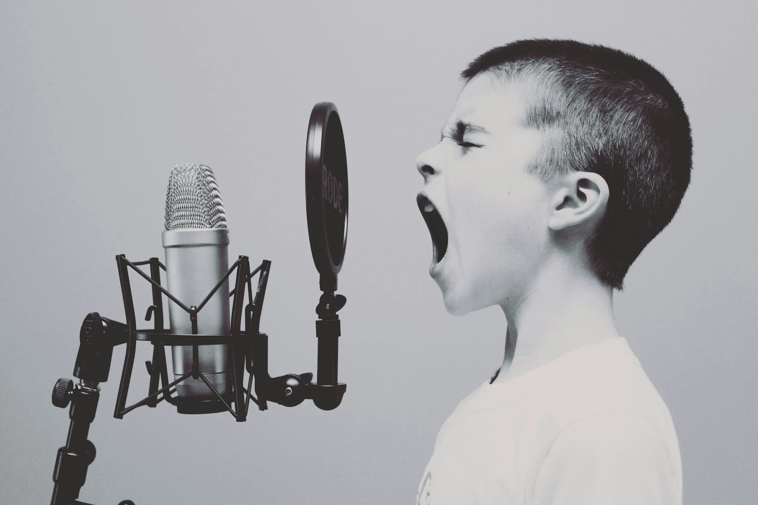 Black and white photo of a young boy with a short haircut singing into a professional microphone with a pop filter, wearing a plain shirt.