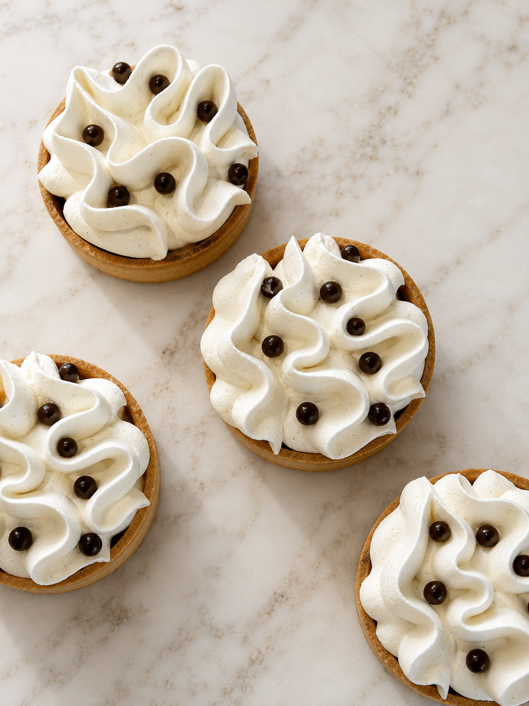 Four tartlets with whipped cream and small dark pearls on a white marble surface.