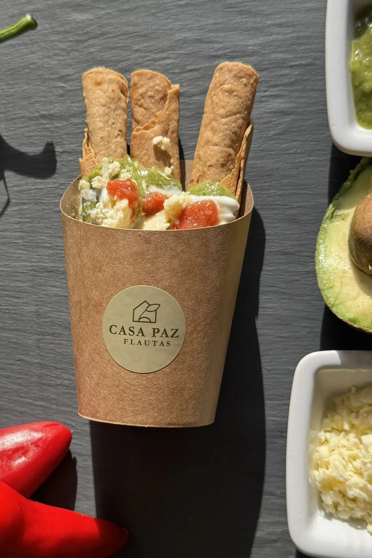 A paper cup with the label 'Casa Paz Flautas' filled with three fried flautas garnished with green sauce, white sauce, and red salsa, on a dark surface with small dishes of green and white toppings nearby.