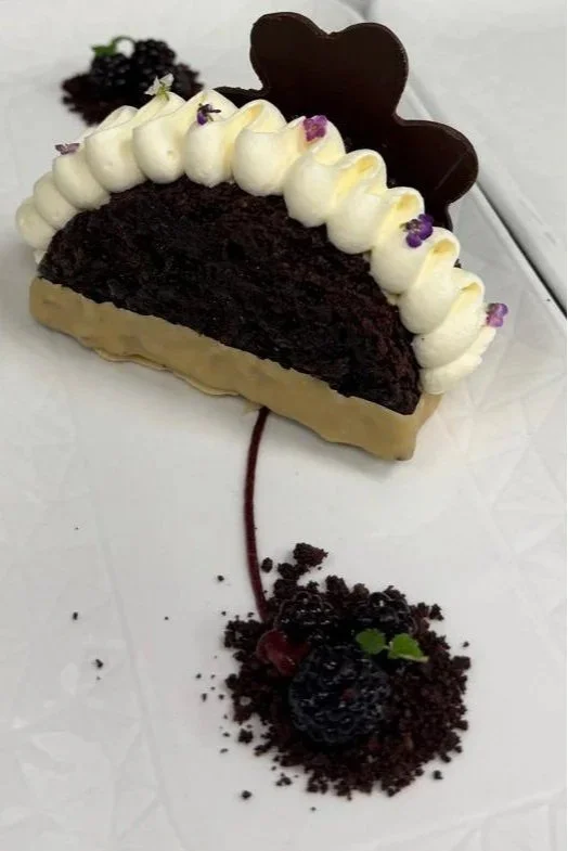 A slice of chocolate cake decorated with whipped cream and small purple edible flowers, with a chocolate heart on top. The cake is placed on a white plate, and a cherry with chocolate crumbs and a small green leaf are arranged in a pattern resembling a cherry stem and cherry on the plate.