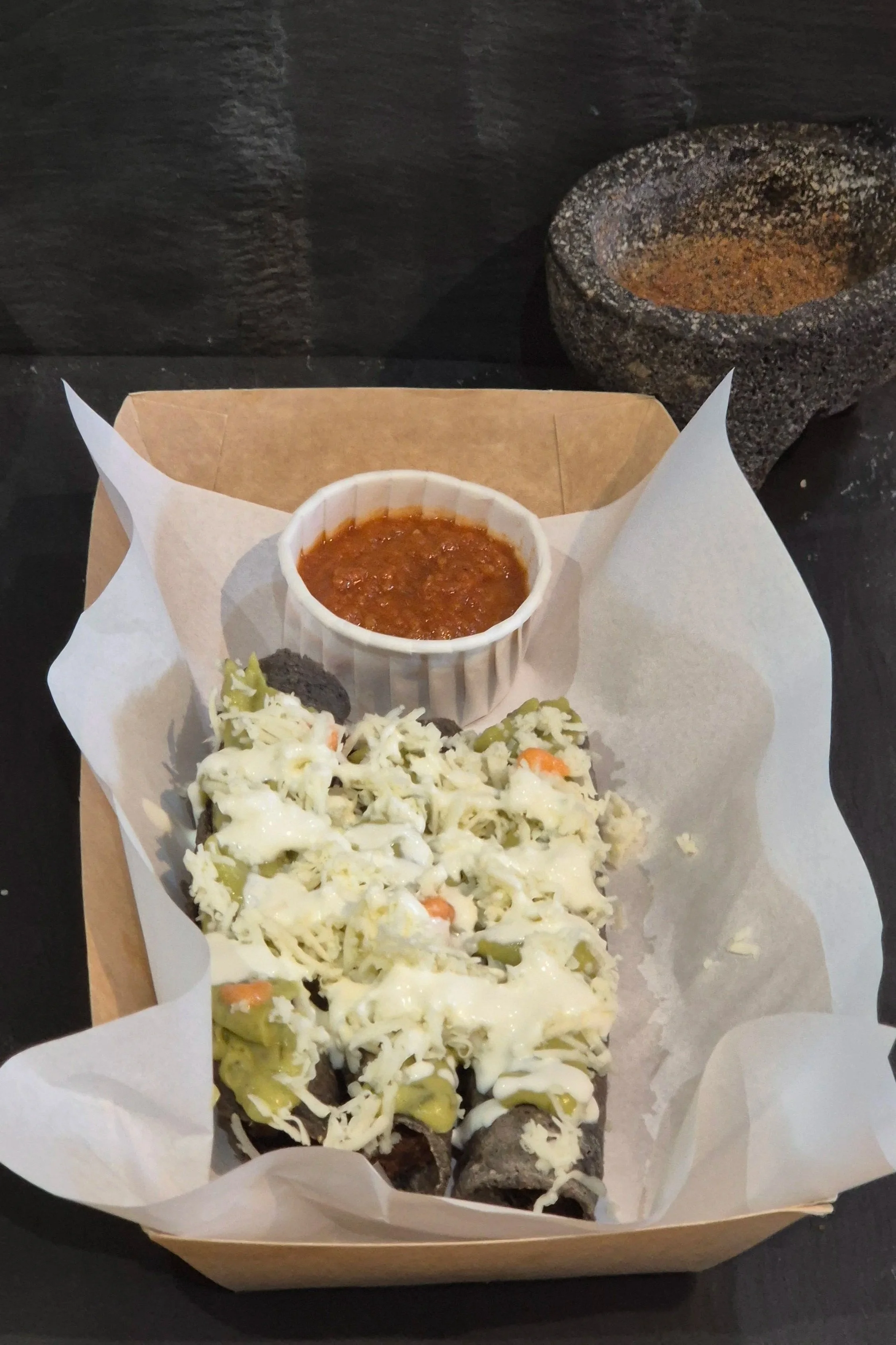 Open cardboard food tray lined with parchment paper containing two mexican Flautas topped with shredded cheese, with a small bowl of red chili sauce. 