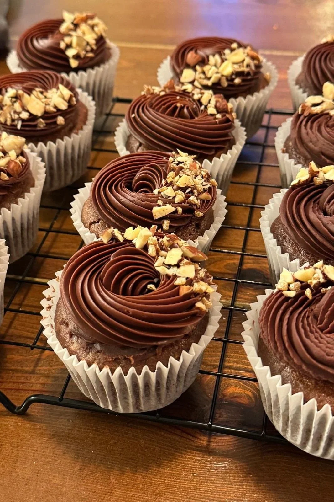 Several chocolate cupcakes with swirled chocolate frosting and chopped nuts on top, cooling on a wire rack.