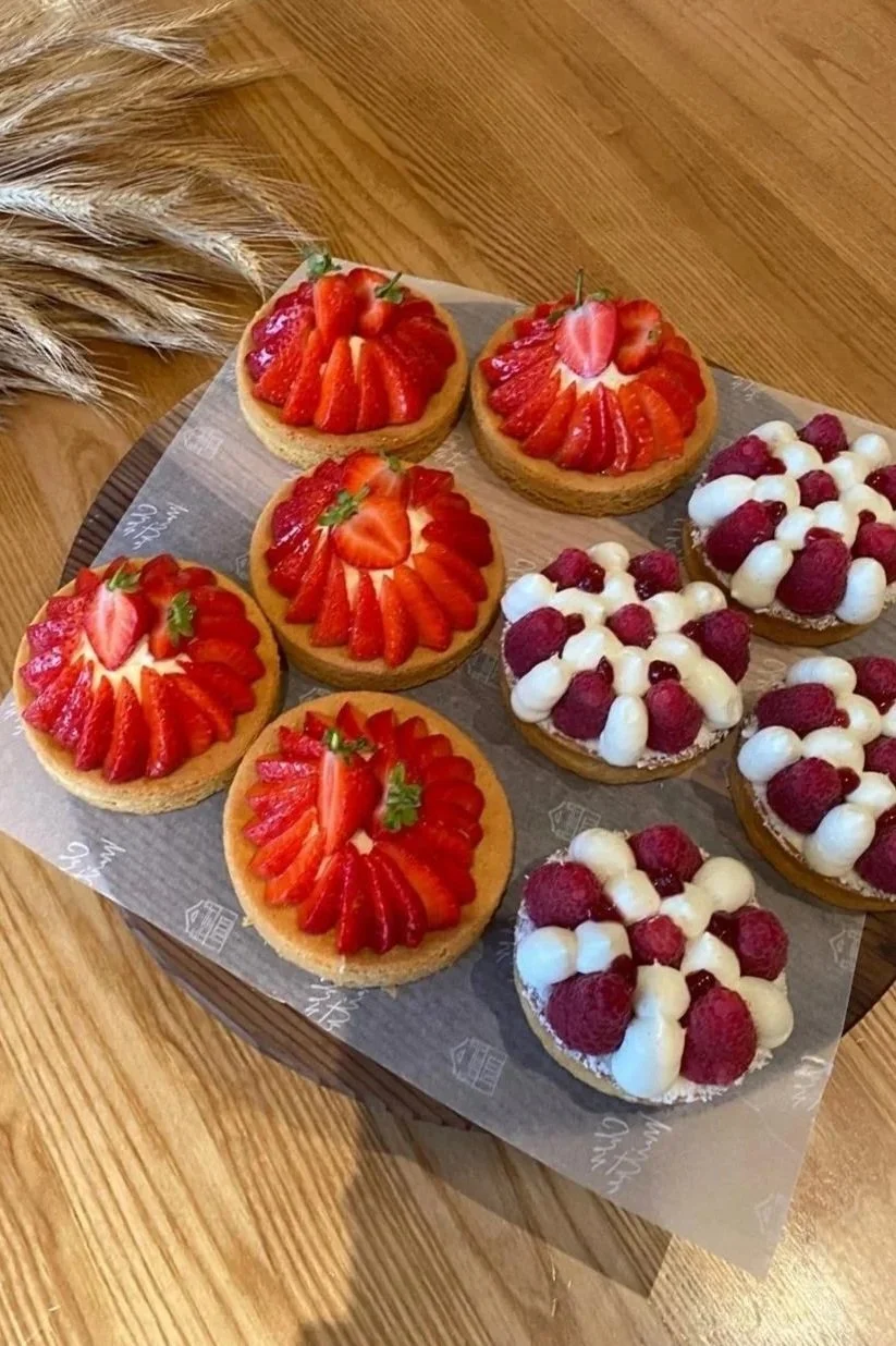 Assorted fruit tarts topped with sliced strawberries and fresh raspberries with cream, displayed on a wooden table.