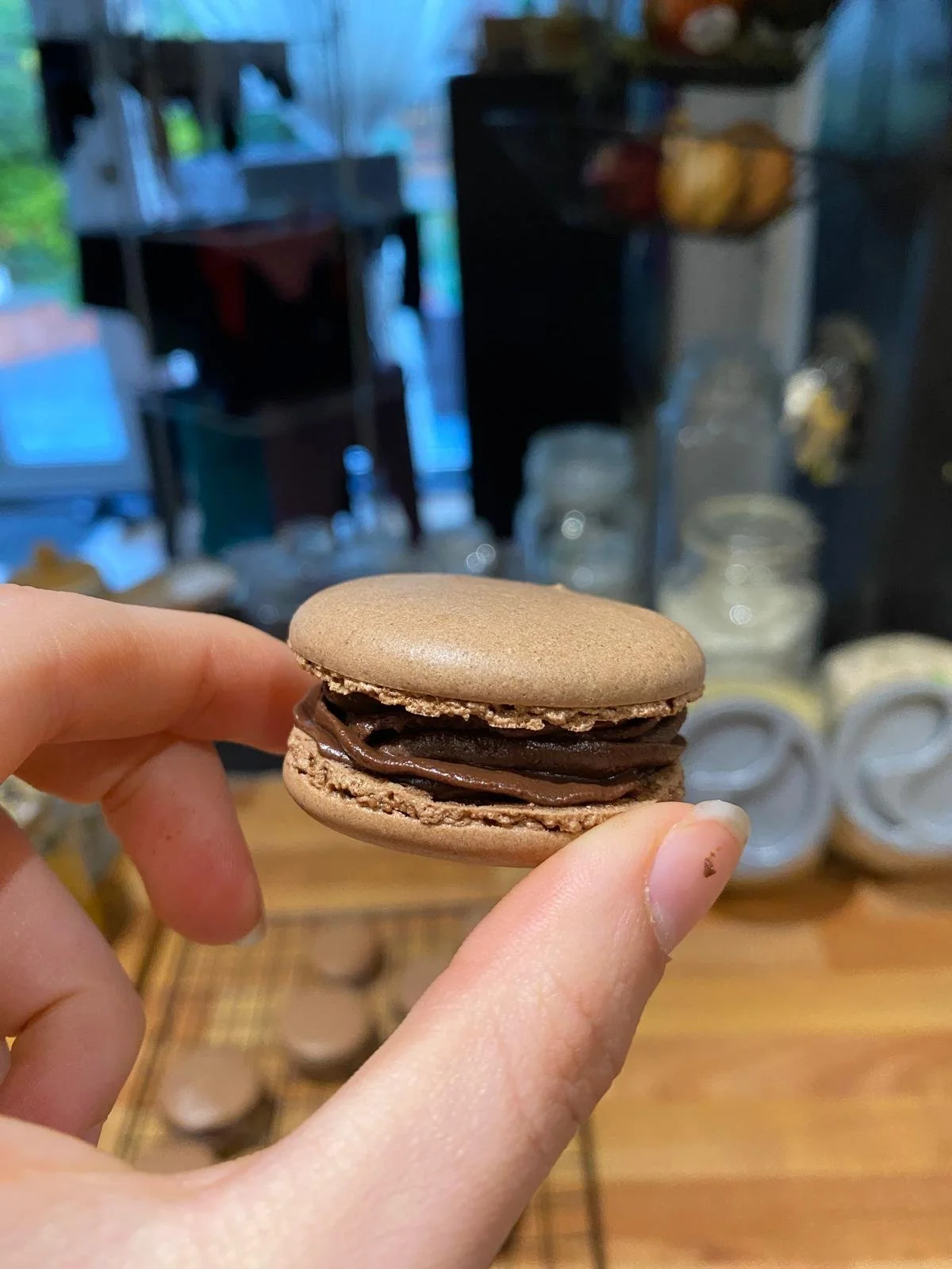 Person holding a chocolate macaron with chocolate filling, blurred background showing shelves with jars and kitchen items.