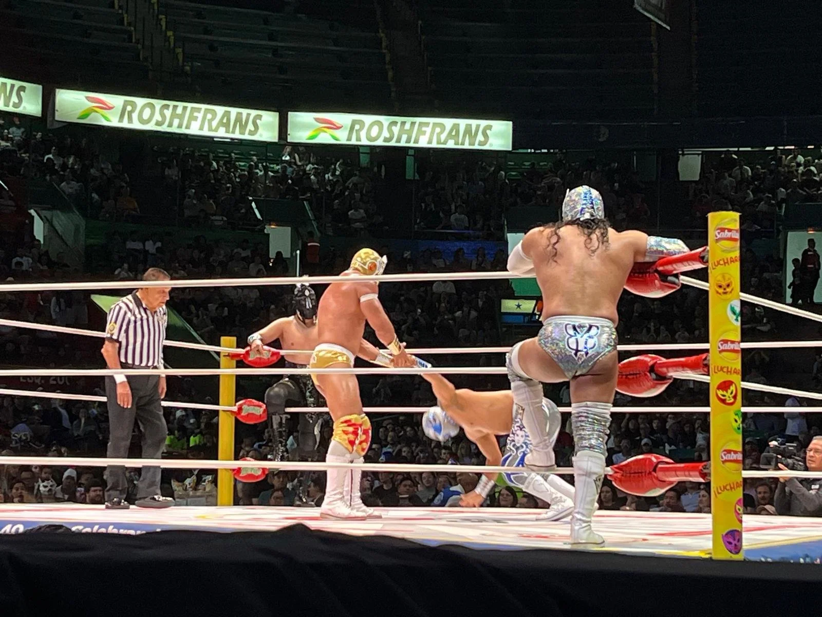 Mexican wrestlers in a lucha libre match inside a wrestling ring, with a referee observing. One wrestler is on his knees, another is standing, and two are on the mat. The background shows a large crowd and advertising banners.