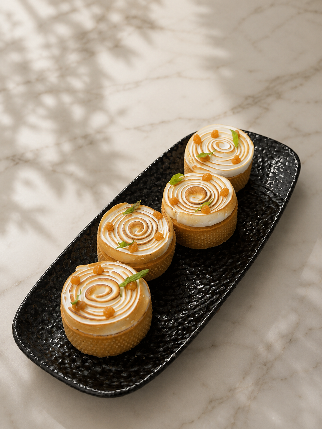Four small, round desserts with swirled white topping, garnished with small green leaves and tiny orange dots, on a black textured rectangular plate on a marble surface.