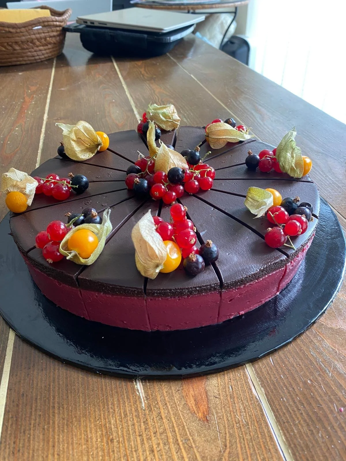 Chocolate cake decorated with red currants, black currants, Cape gooseberries, and physalis on top, sliced into pieces, on a black serving board on a wooden table.