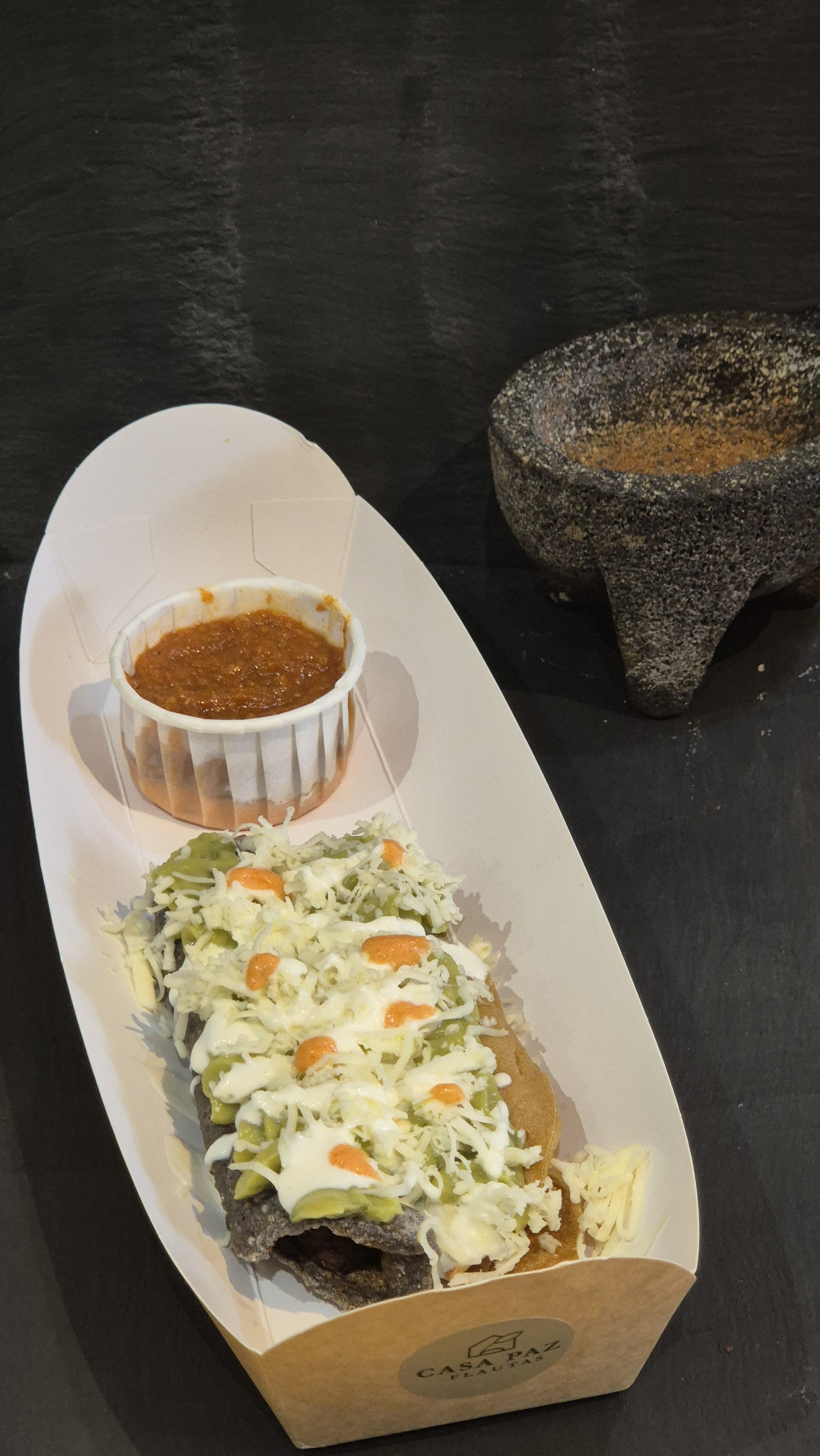 A cardboard tray holding Mexican flautas,  topped with cheese, guacamole, and hot sauce, with a small container of chili and a bowl of spice on a black surface.