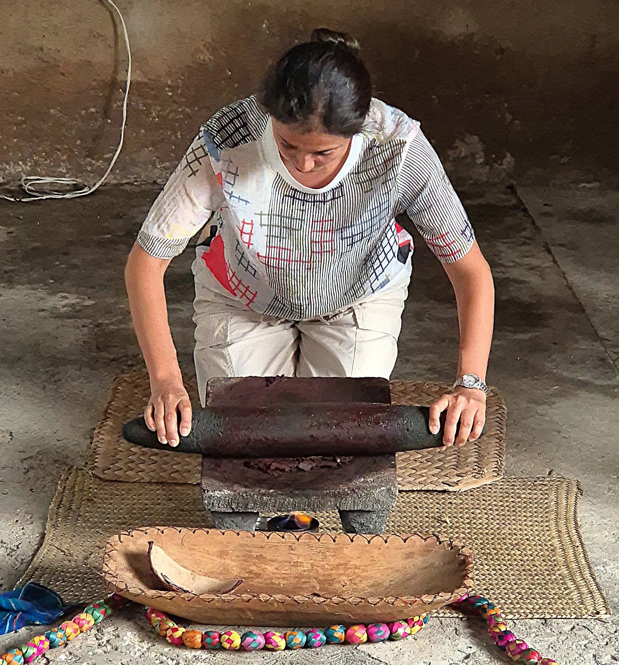 Woman kneeling on ground, working on a colorful, woven rope border, with a carved wooden structure in front of her, in an indoor setting with a concrete or dirt floor and a brown textured wall.