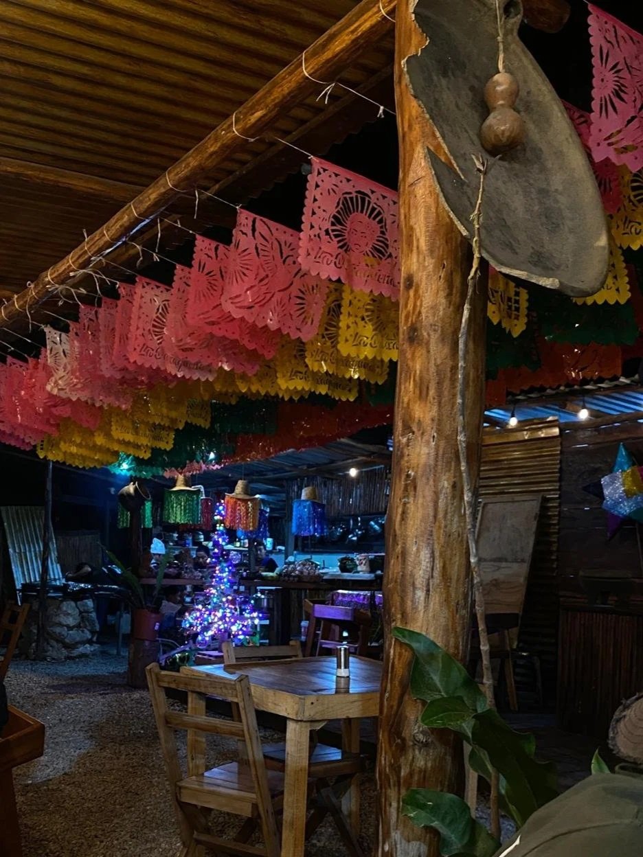 Decorative paper banners hanging from a wooden ceiling in a rustic, festive setting with colorful lights, a small Christmas tree, and wooden furniture.