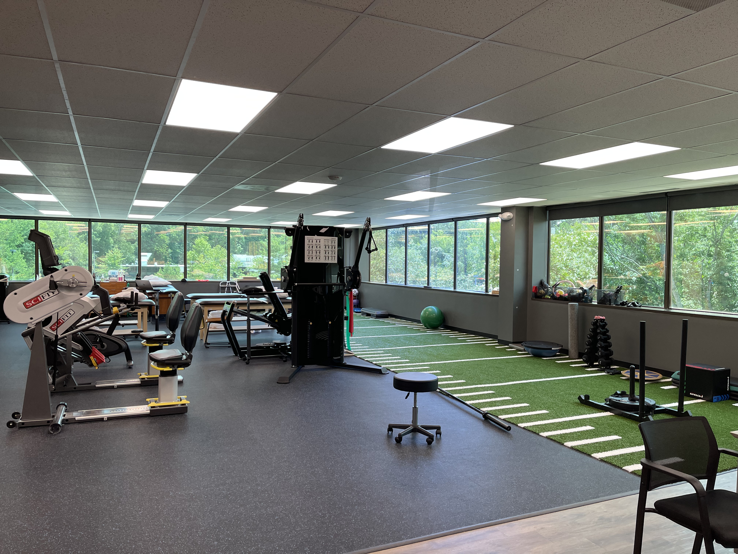 An indoor fitness and recovery center with exercise equipment, a green turf area, large windows showing trees outside, and bright overhead lighting.