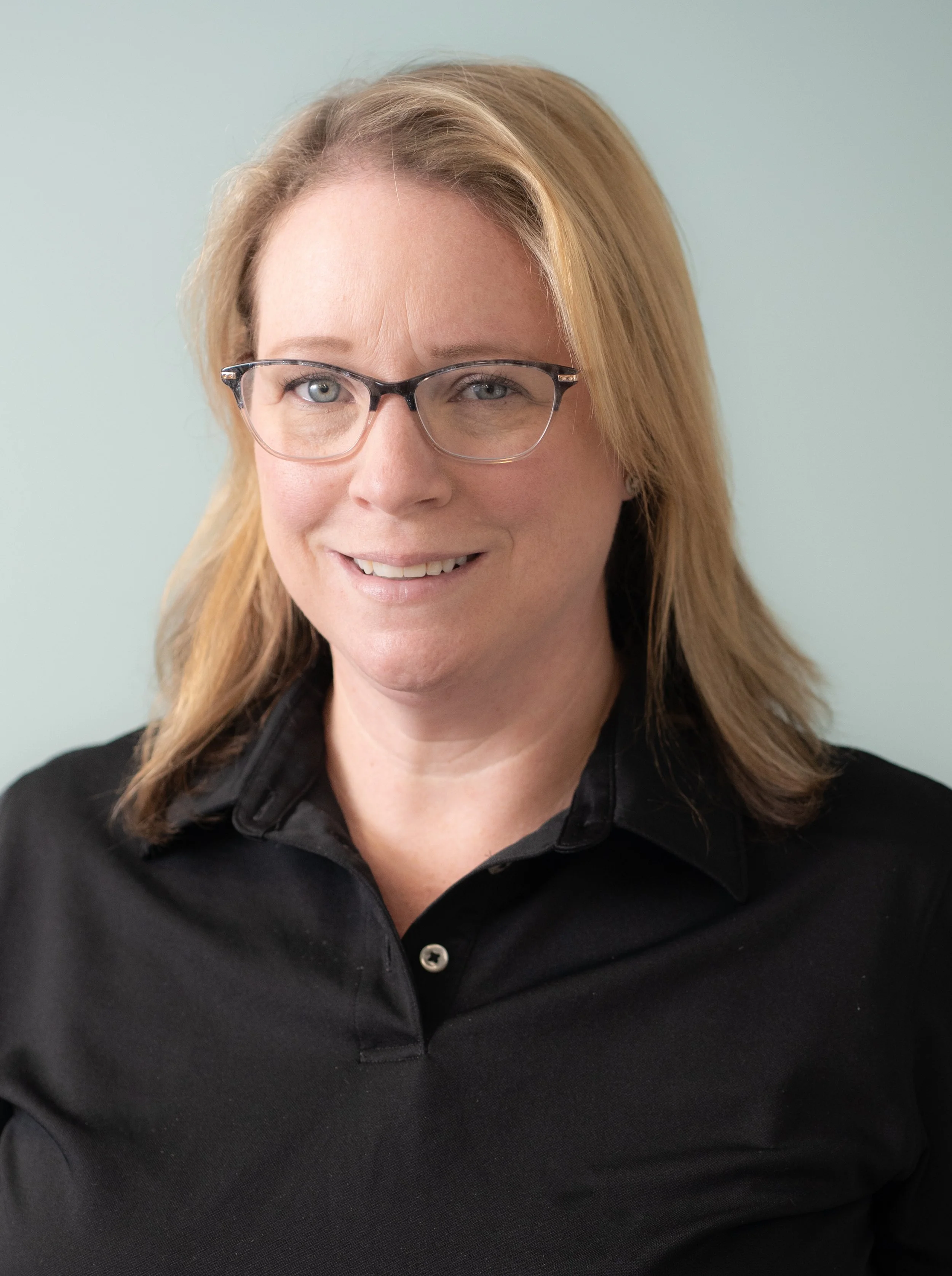 A woman with light skin, shoulder-length light brown hair, and glasses, smiling while wearing a black shirt against a plain, light-colored background.