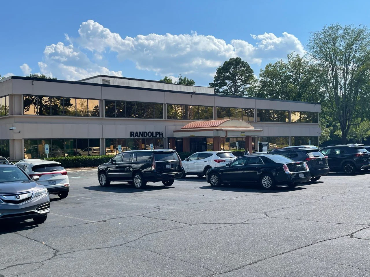 Parking lot with multiple cars in front of a two-story office building labeled 'RANDOM.' The building has large windows, a curved awning over the entrance, and is surrounded by trees under a partly cloudy sky.