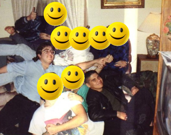 A group of seven people in a cozy living room, some sitting on a couch and some lying down, with yellow smiley face emojis covering their faces. The room has a lamp, a side table, and a framed picture on the wall.