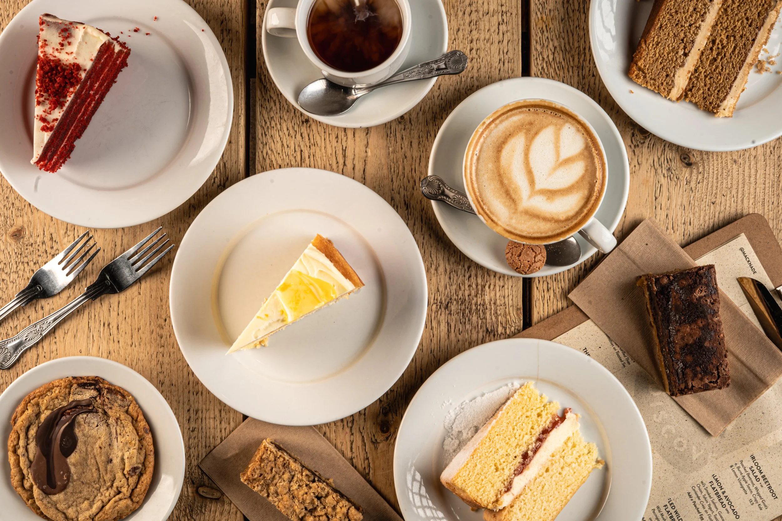 Assorted desserts and coffee on a wooden table, including a slice of red velvet cake, a slice of lemon cheesecake, a chocolate chip cookie, a piece of banana cake, and a cappuccino with latte art.