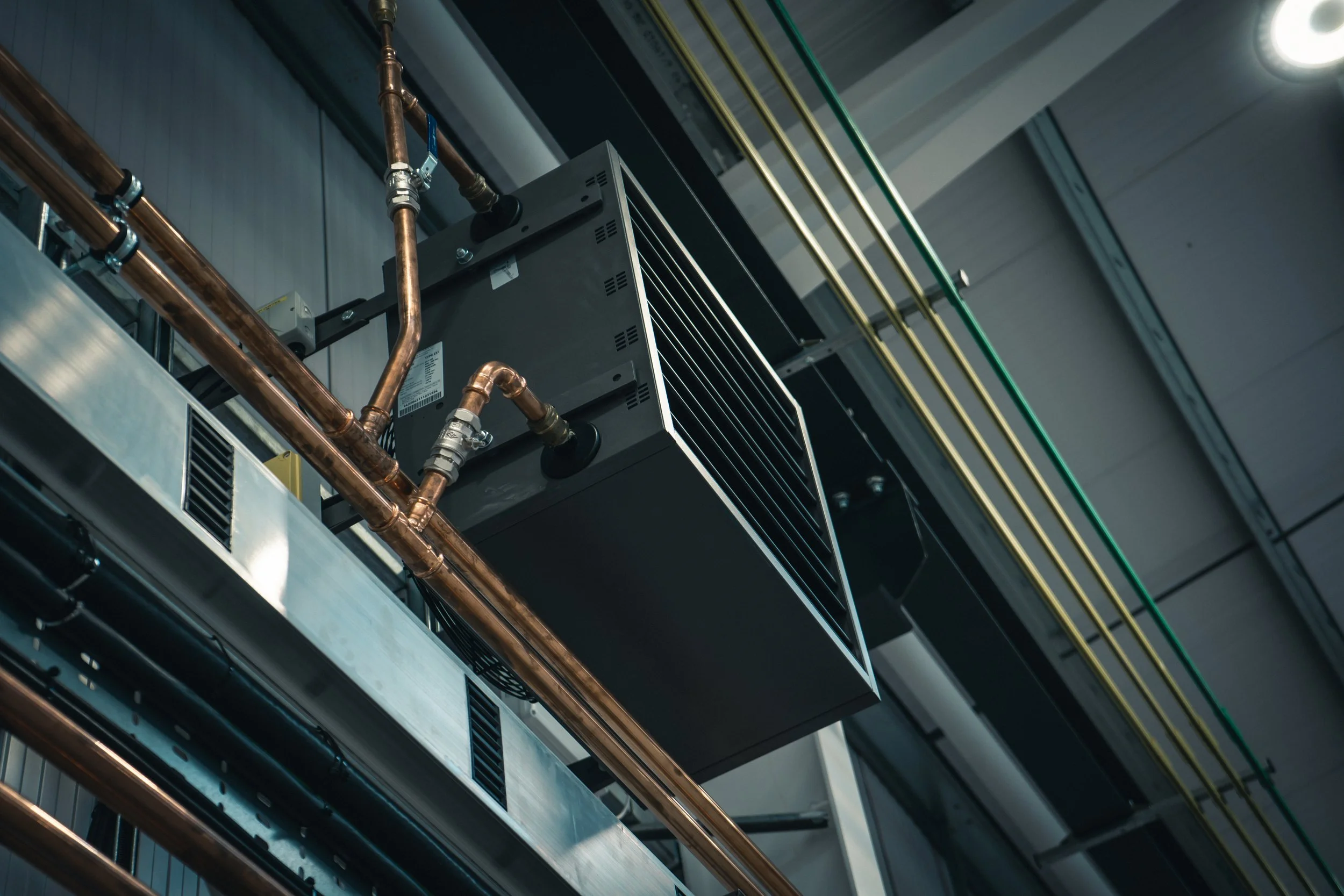 View of an industrial HVAC system featuring copper pipes, metal boxes, and various wiring, mounted on a ceiling in a commercial or industrial building.