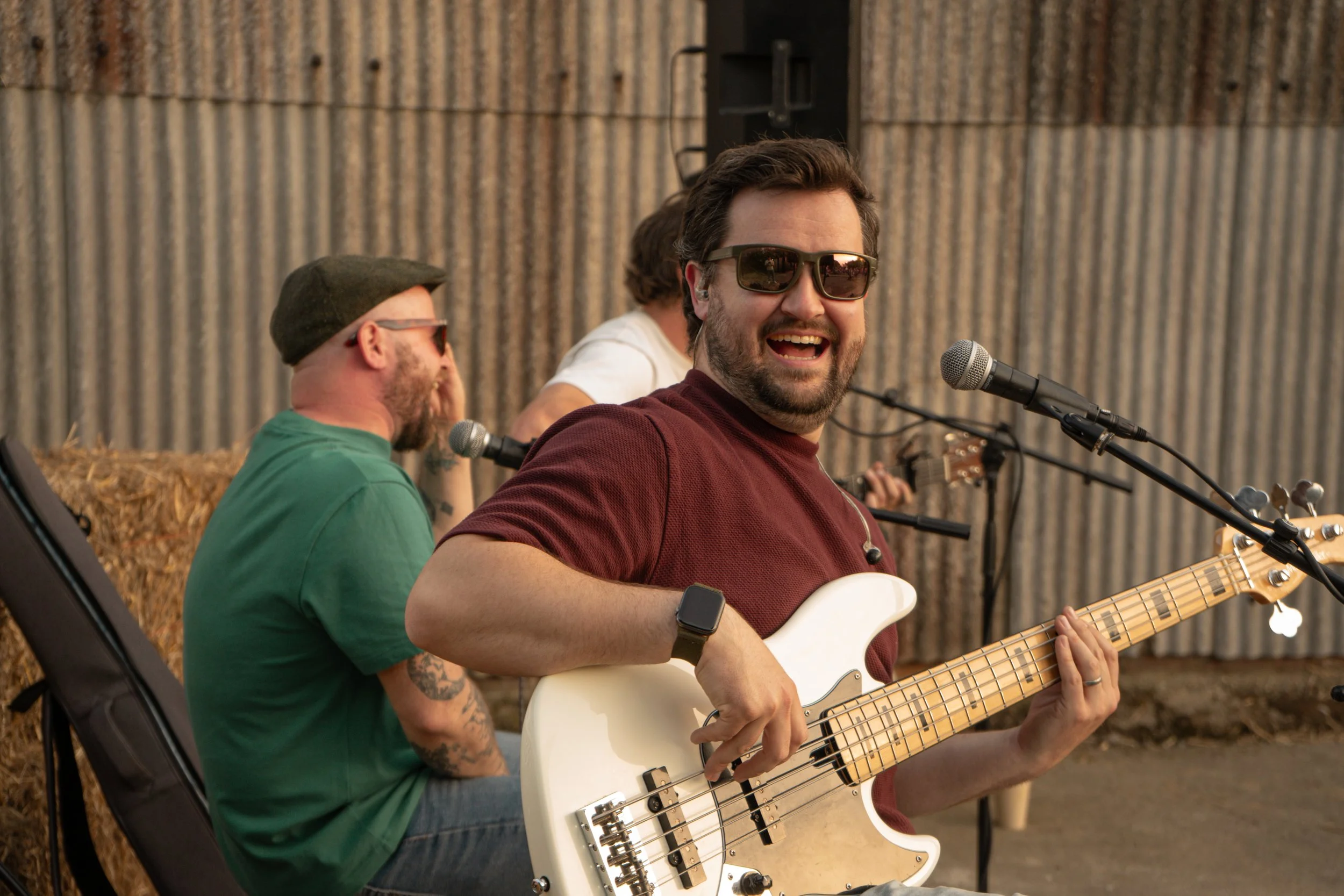 Three men playing guitars outdoors against a corrugated metal wall, smiling, wearing sunglasses.