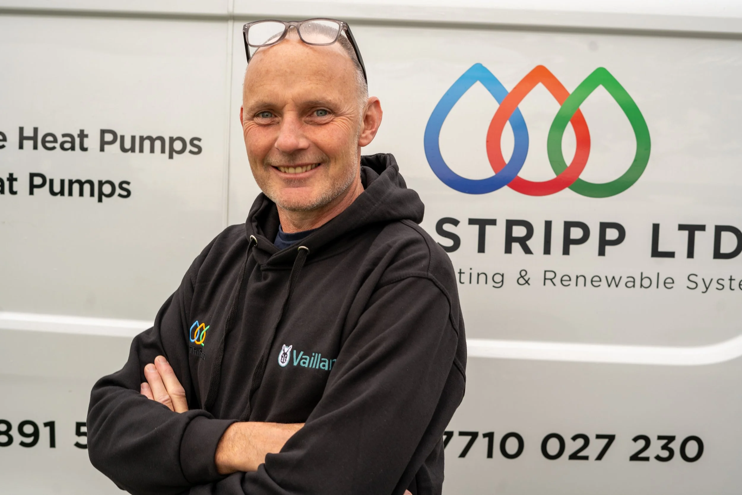 A man with glasses on his head, smiling with arms crossed, standing in front of a van with colorful logo and text related to heating and renewable systems.