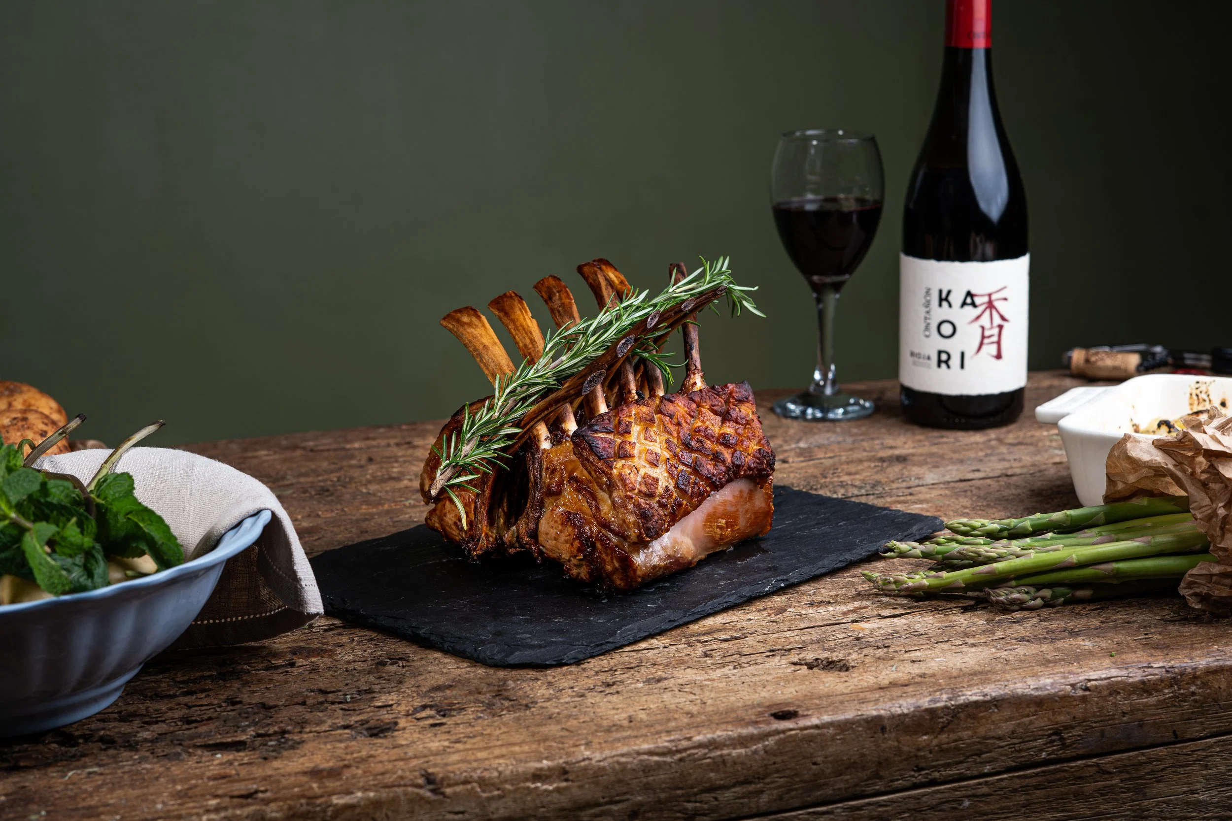 A roasted rack of lamb garnished with rosemary on a dark slate serving board on a rustic wooden table, with a bottle of red wine, a filled wine glass, and various vegetables and herbs in the background.