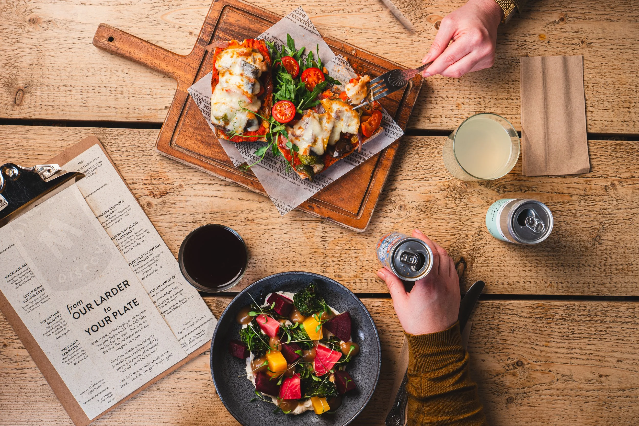 A wooden table with a cutting board holding a slice of pizza topped with cheese, vegetables, and potentially cheese or sauce. There is a salad with cherry tomatoes, mixed greens, and vegetables, and a glass of lemonade. A person is holding a fork wit