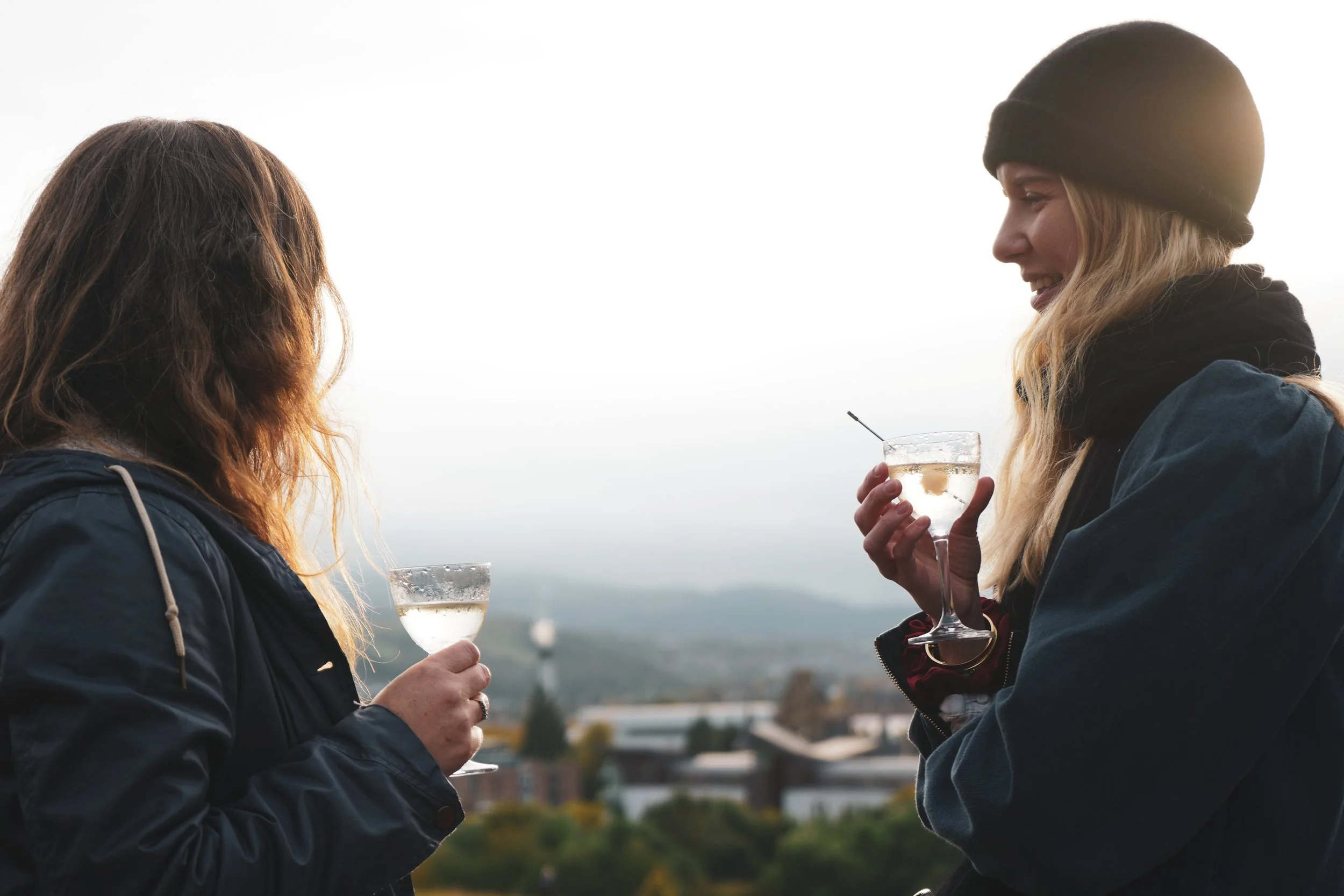 Two women holding glasses of white wine and engaging in conversation outdoors, with a blurry cityscape and trees in the background during sunset.