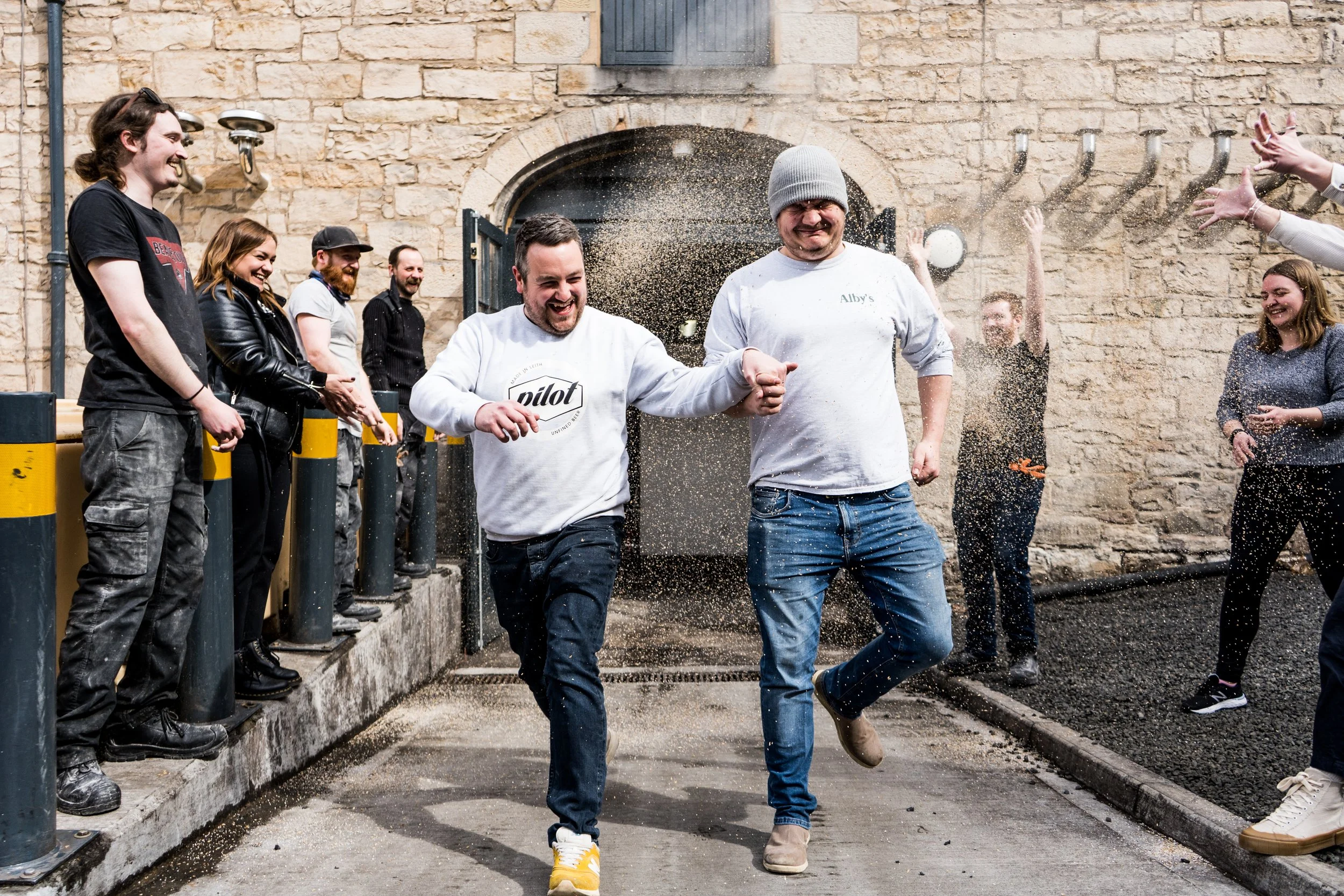 A group of people celebrating outside near a stone building, with one person in a gray beanie and another in a white sweatshirt holding hands and running through a pile of confetti or flour, smiling and laughing.