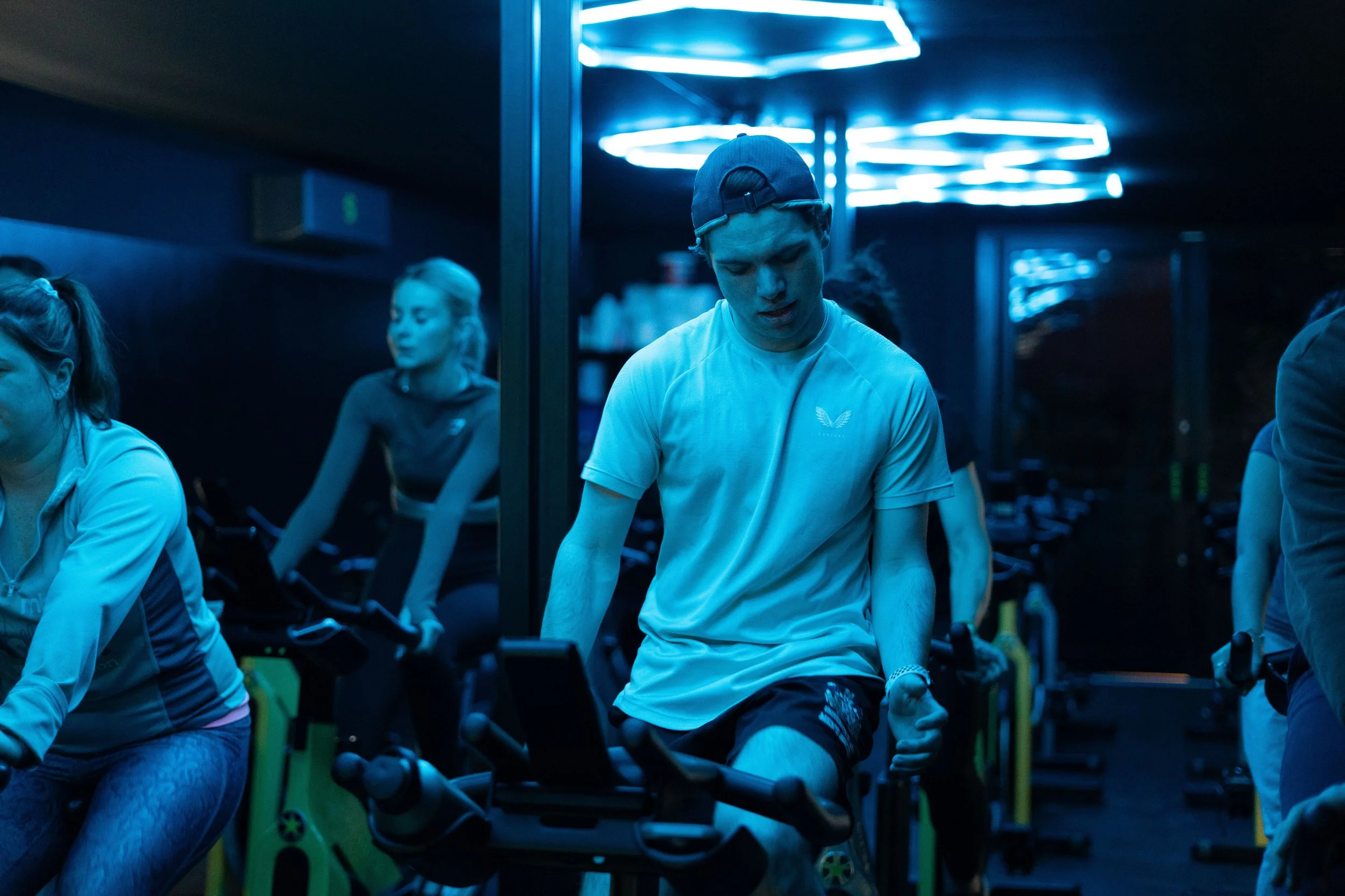 People exercising on stationary bikes in a dimly lit gym with blue neon lighting.