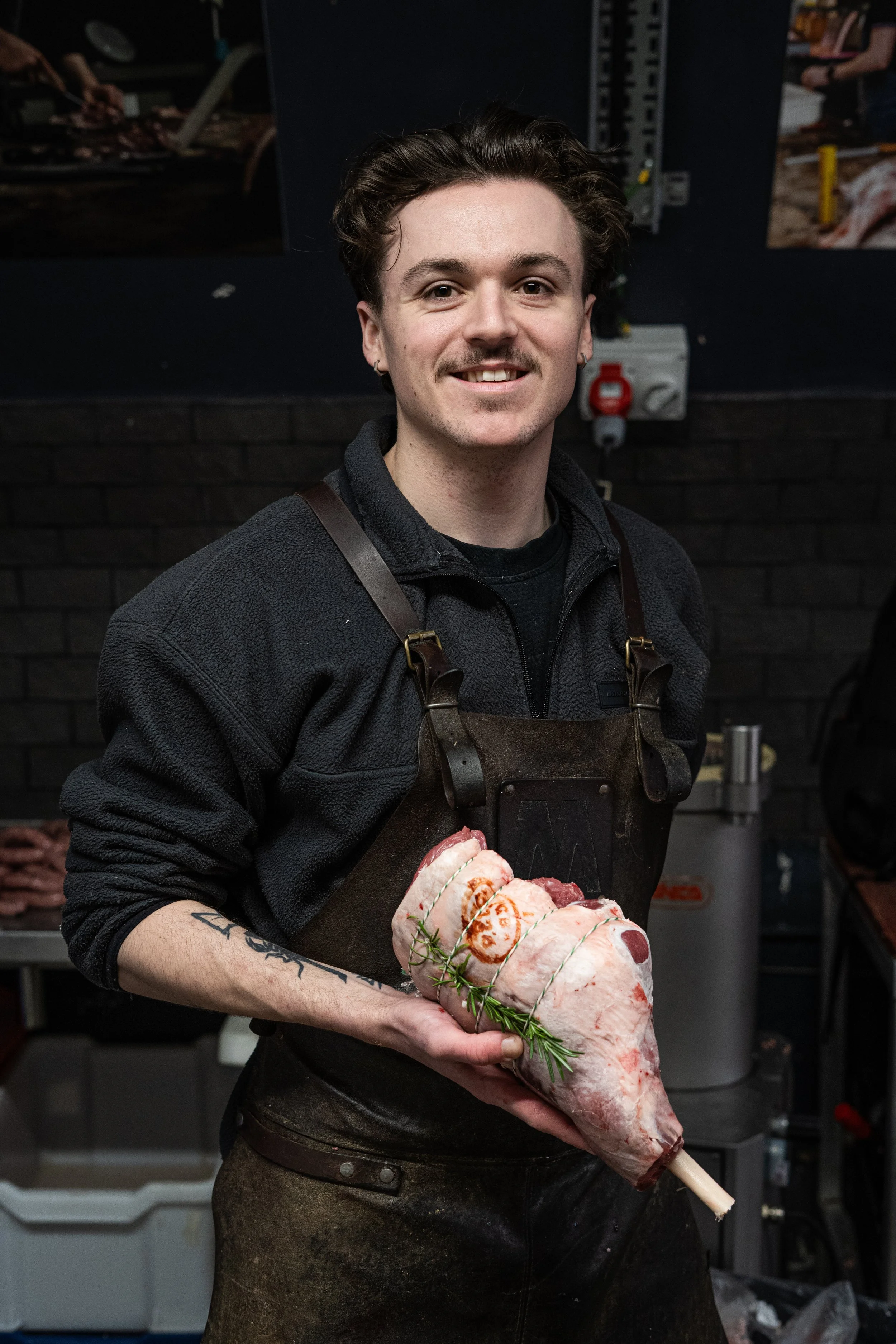 A young man with brown hair and earrings, wearing a black fleece jacket and a leather apron, is holding a large raw pig's leg in a butcher's shop or restaurant kitchen, with meat and equipment in the background.