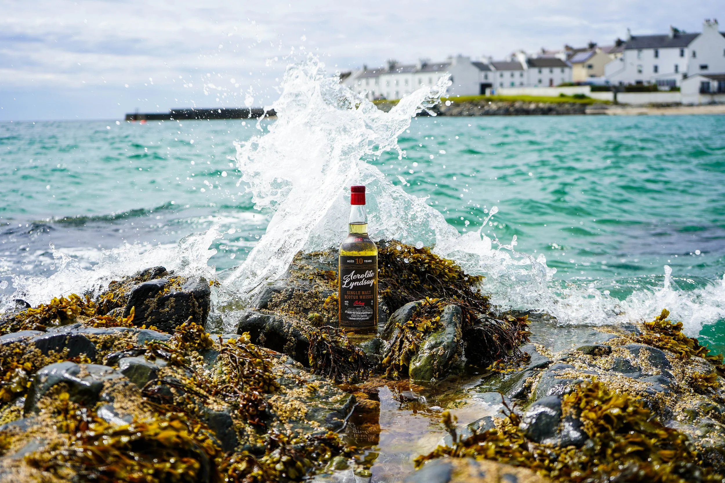 A bottle of Aerolite Lyndsay single malt Scotch whiskey placed on rocks on a shoreline with seaweed, with water splashing around it. In the background, there are houses along the coastline under a cloudy sky.