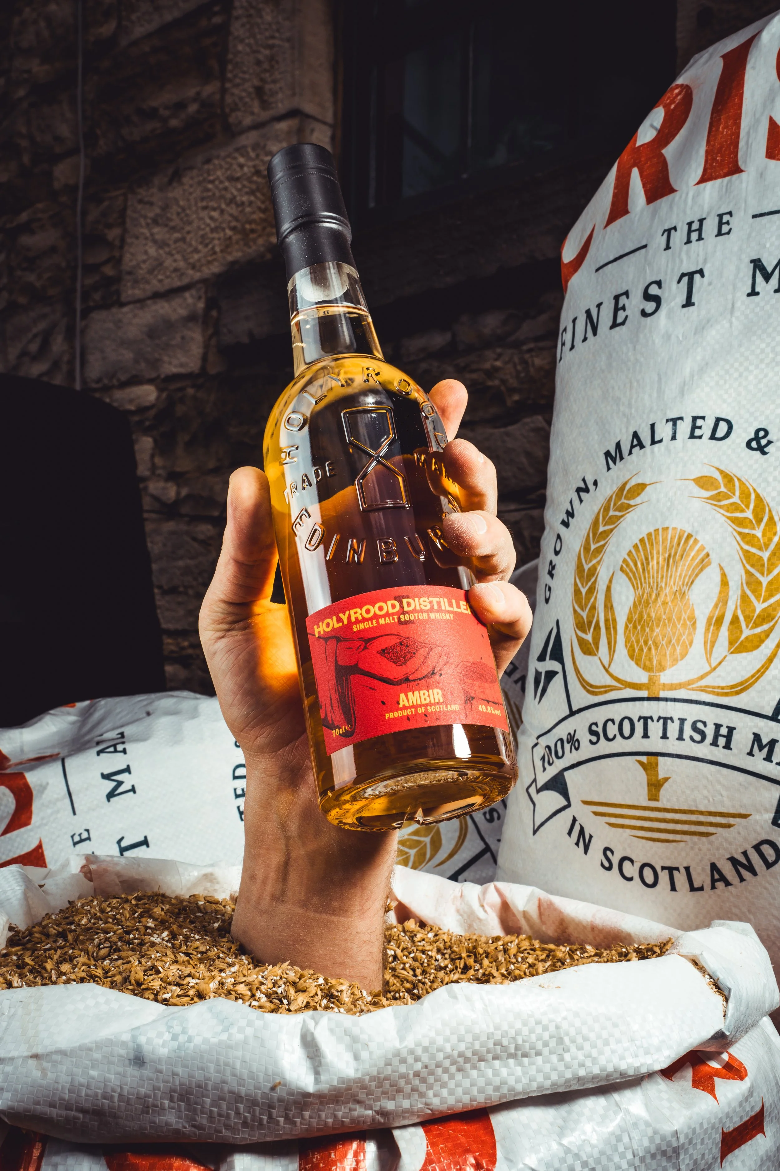 A person holding a bottle of Holyrood Distillery single malt Scotch whisky over a large bag of crushed malted barley, with a stone wall in the background.