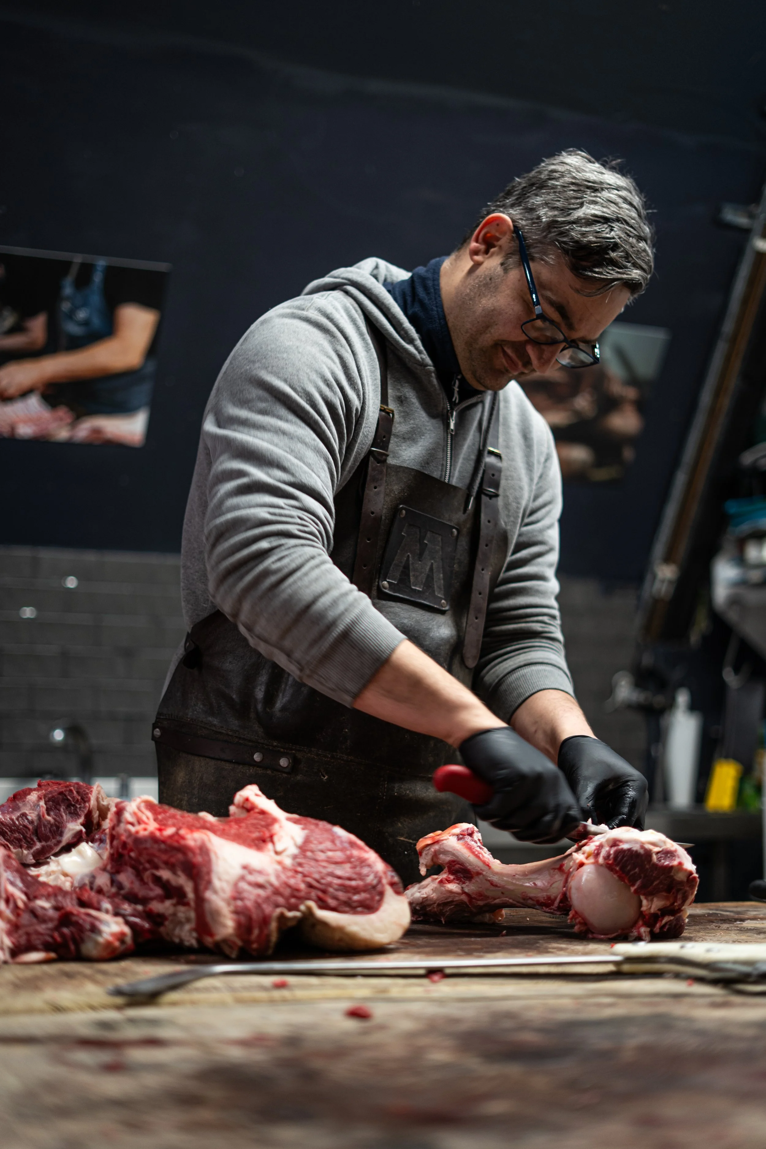 A man with glasses and a gray hoodie, wearing a black apron, butchering large cuts of raw beef on a wooden cutting board in a meat processing room with dark walls and framed pictures in the background.