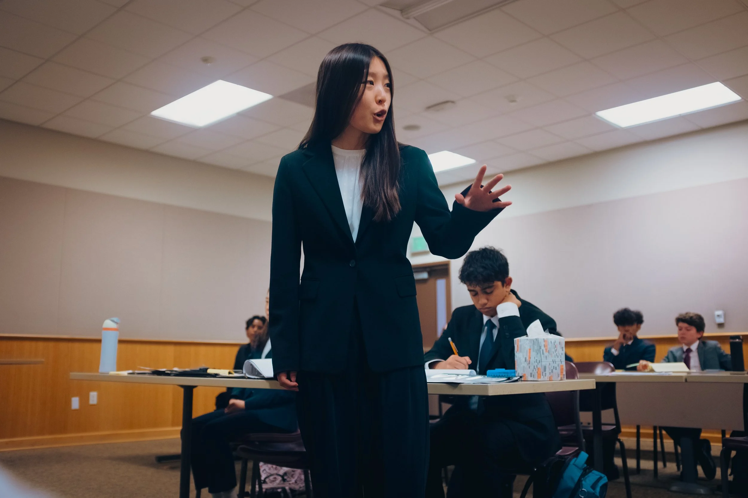 A woman in a dark blazer and white shirt is standing in a classroom, giving a closing argument in a high school mock trial competition. The SoCal Mock Trial League hosts high school mock trial tournaments across Southern California.