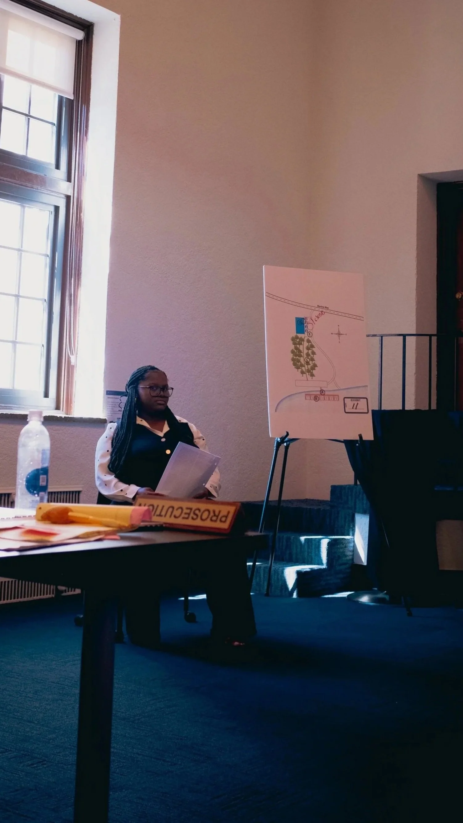 A high school student sits in a chair, acting as a witness in a high school mock trial tournament. She sits beside a demostrative on a podium. The SoCal Mock Trial League hosts high school mock trial tournaments across Southern California.