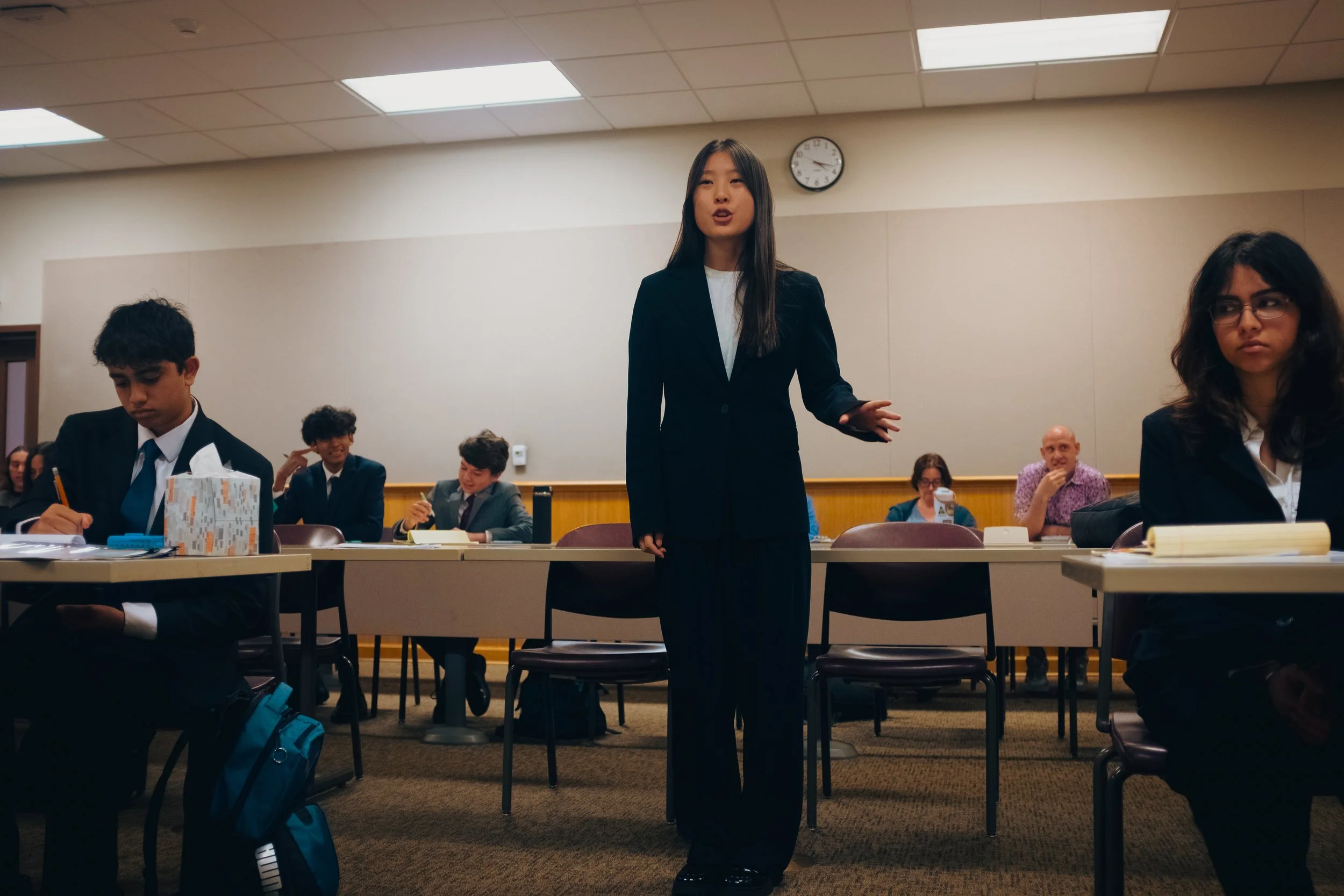 A high school student in a black blazer and white shirt is standing between council tables, presenting a statement. The Southern California Mock Trial League hosts high school mock trial tournaments across Southern California.