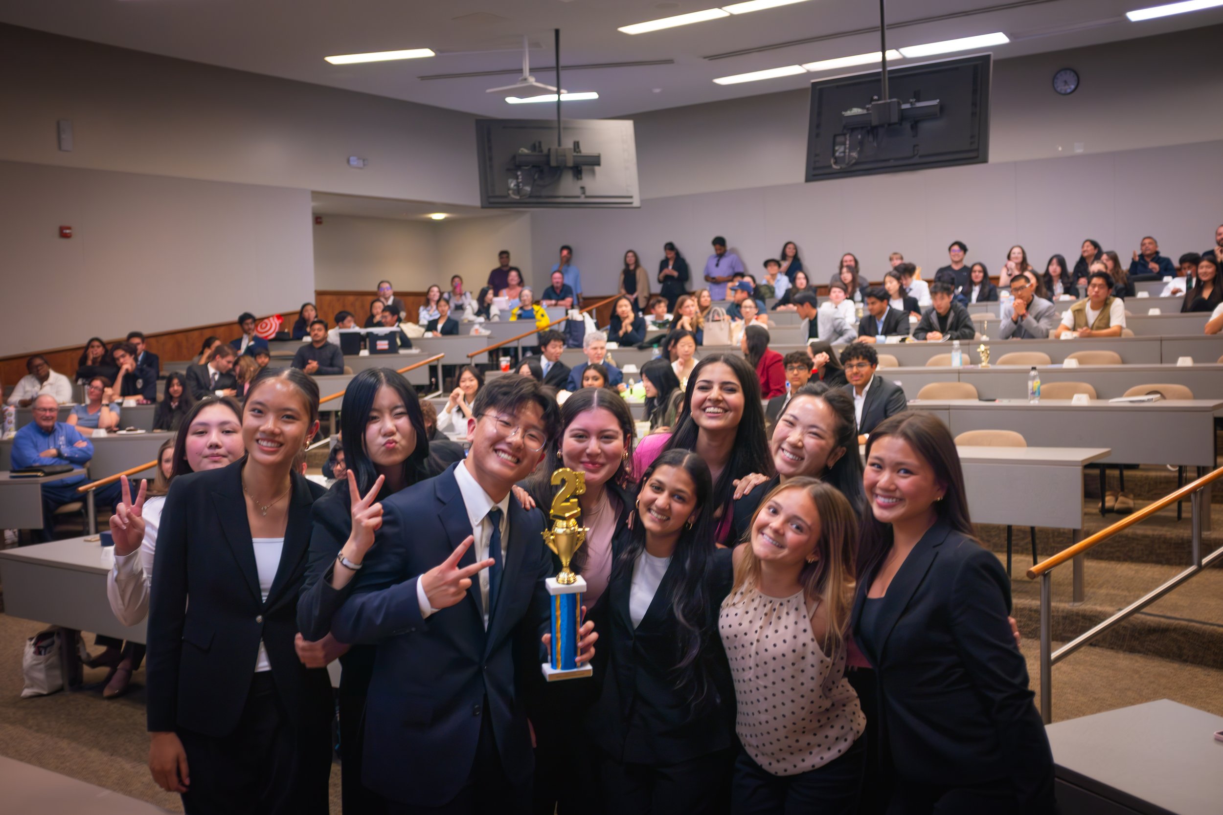 Group of smiling high school students in business attire celebrating with a trophy after winning second place at a high school mock trial tournament. The SoCal Mock Trial League hosts high school mock trial tournaments across Southern California.